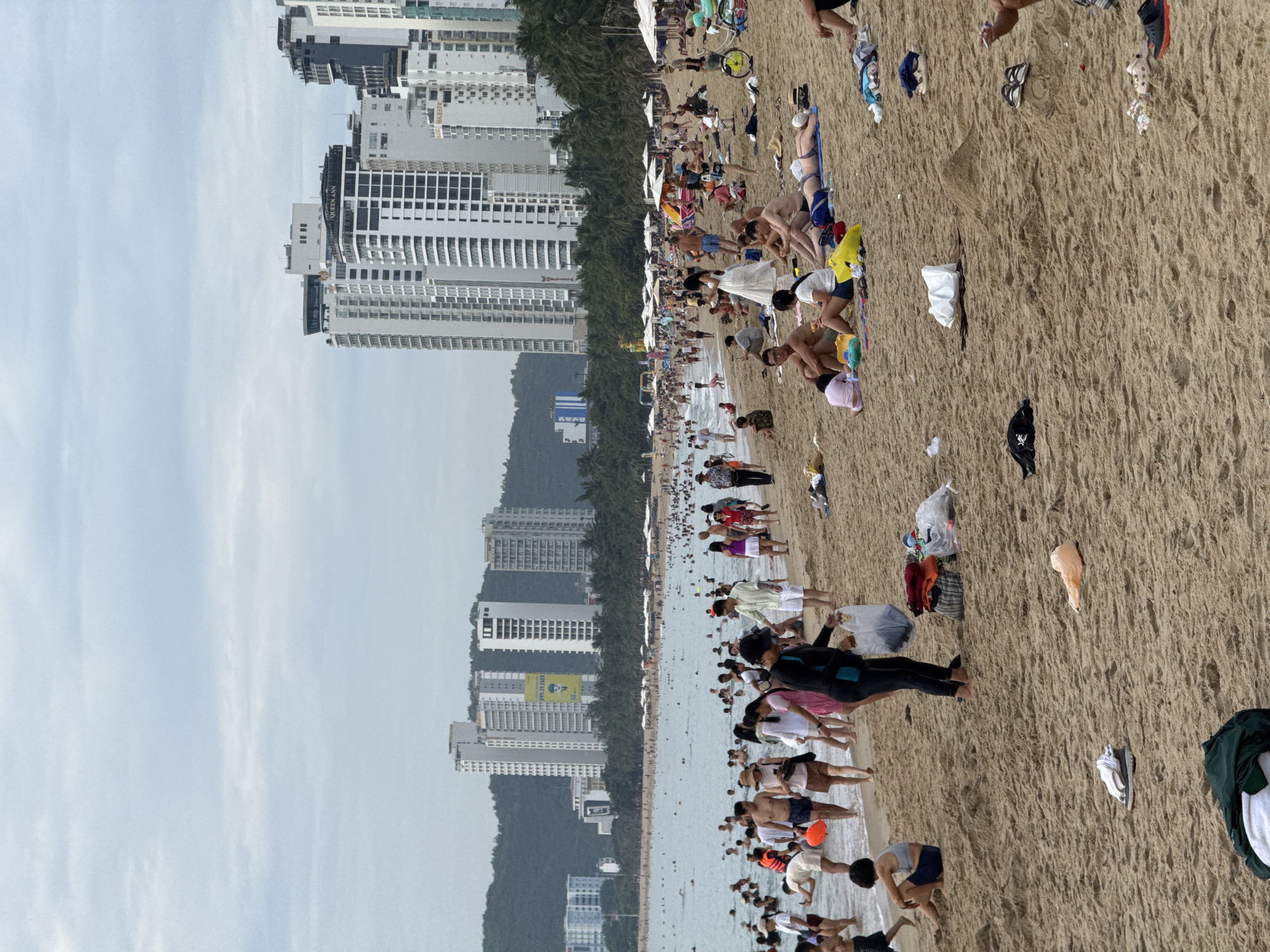 The image depicts a crowded beach scene in Nha Trang, Vietnam, with a backdrop of modern high-rise buildings and lush green hills. The sandy beach is populated by numerous individuals engaged in various beach activities. In the foreground, people are seen sitting, walking, and lying on the sand, some in swimwear and others in casual attire. Several piles of clothing and bags are scattered on the beach, indicating people's belongings.
The water is filled with bathers, with some closer to the shore and others further out. Umbrellas are set up along the beach, providing shade for beachgoers. In the background, a dense line of palm trees separates the beach from the towering hotels and apartment buildings that characterize Nha Trang's coastline. The sky is overcast, suggesting a cloudy day. The overall impression is a lively and bustling beach atmosphere, a popular destination for both locals and tourists.