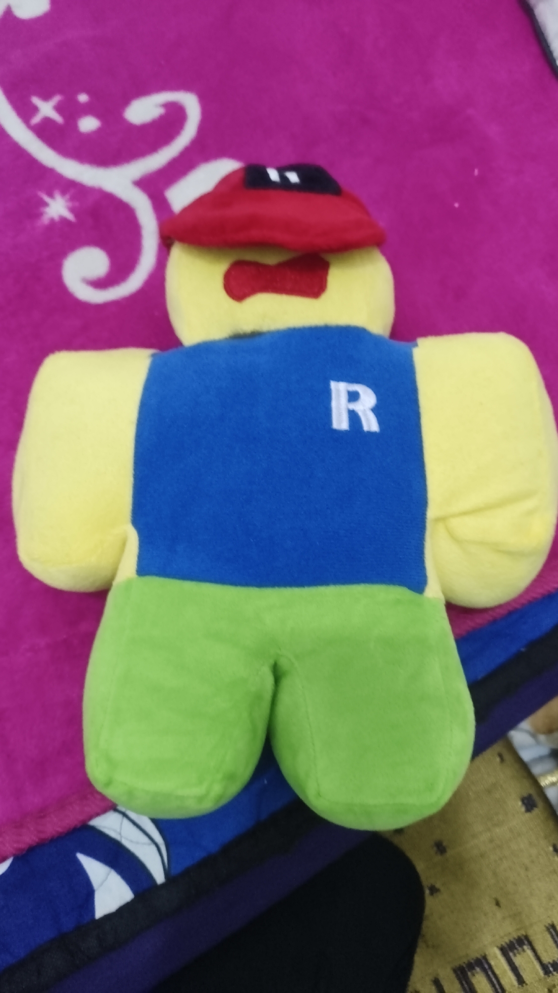 A Roblox Noob plush toy is positioned upright on a pink blanket with a white swirl pattern. The toy has a yellow head with a red hat featuring black squares and a red bow tie. Its body is blue with a white "R" on the back, and its legs are green. The background consists of the pink blanket and a dark surface below. This image was taken in Betung, Indonesia.