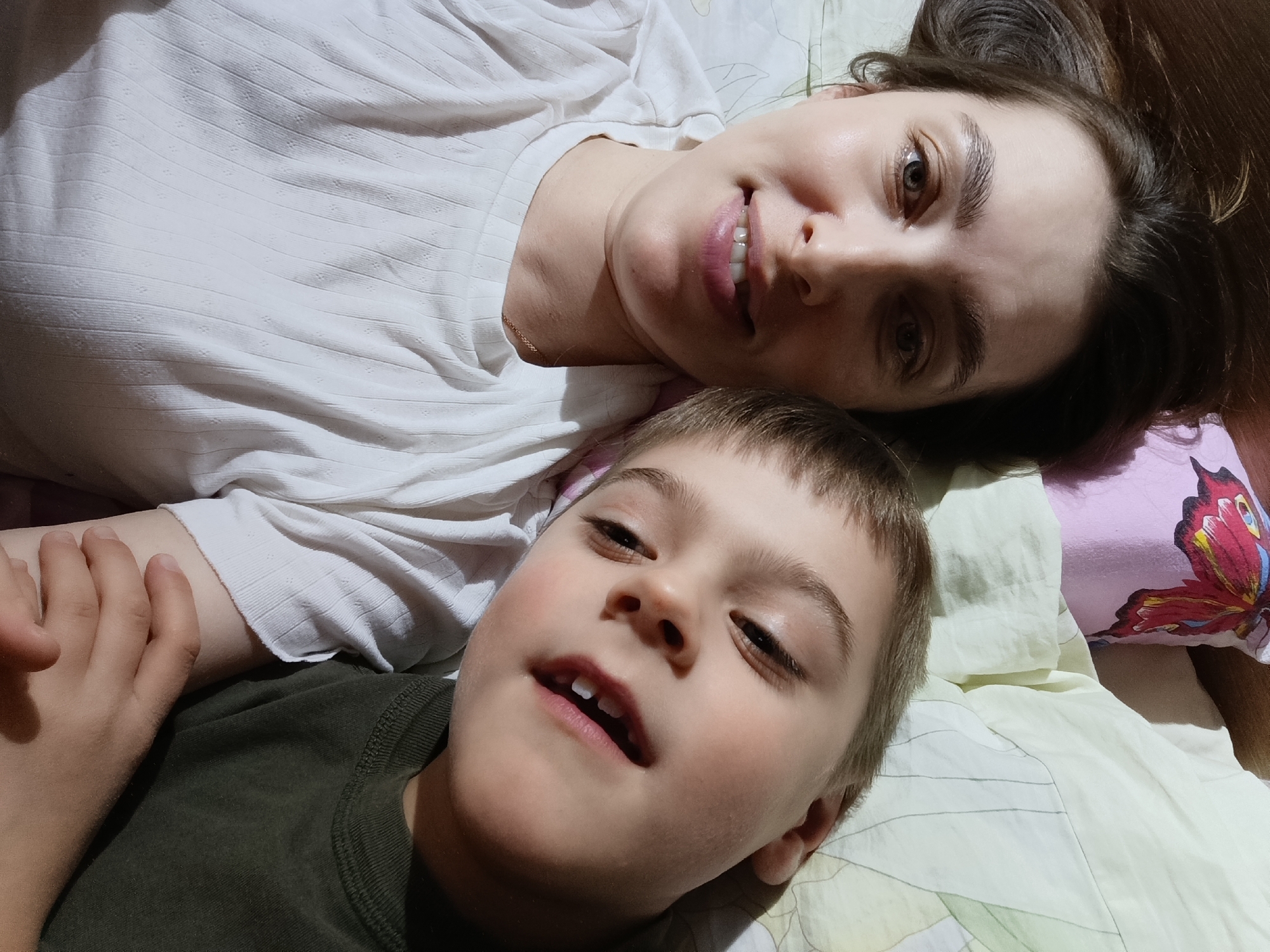 Two individuals, likely a mother and child, are lying on a bed in an indoor setting. The woman, wearing a white top, is positioned towards the top of the frame, smiling. The child, wearing a dark green shirt, lies below her, looking upwards. The bed is covered with patterned sheets, including one with a butterfly design. The location is in the city of Chaikovsky, Russia.