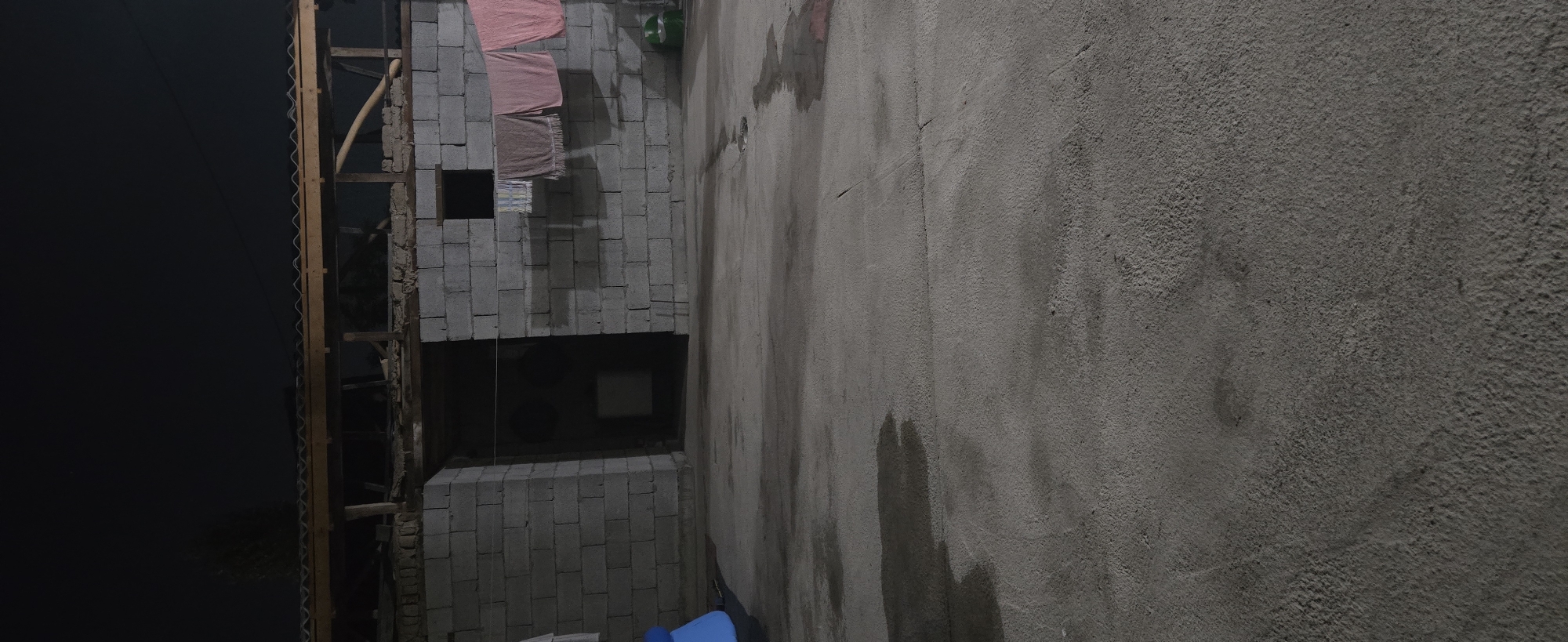 The image depicts an outdoor scene at night in Kurgantepa, Uzbekistan. The primary subjects are a partially constructed building made of concrete blocks and a textured concrete wall. On the side of the block building, several pieces of fabric, possibly towels or clothes, are hanging out to dry. There's a dark, rectangular opening in the block wall, suggesting a window or doorway, and a smaller, lighter-colored rectangle within that opening.
The overall setting appears to be a dimly lit outdoor space, likely a yard or alleyway adjacent to the building. The concrete wall on the right side of the image shows patches of dampness, possibly from recent rain or watering. The sky on the left is almost entirely black, indicating the late hour. Wooden beams and supports are visible on the left side of the block structure, hinting at ongoing construction or scaffolding. A green object, possibly a bucket or container, is affixed to the upper part of the block wall.
There is no visible text in the image. The scene conveys a sense of utilitarian simplicity and possibly a lived-in environment, with the drying laundry suggesting human presence. The time of day is clearly night. The weather is not explicitly indicated beyond the damp patches on the wall. The location cues (Kurgantepa, Uzbekistan) are provided externally.