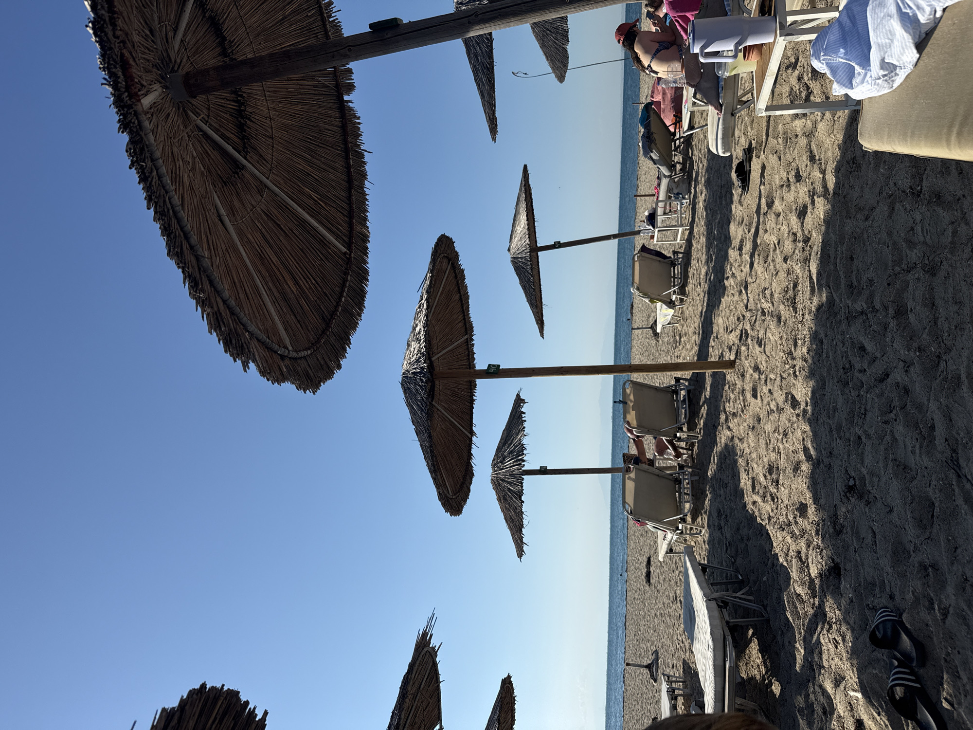 The image depicts a sandy beach on a clear day, likely in Angelochori, Greece. The foreground is dominated by sand and several thatched umbrellas providing shade. In the mid-ground, lounge chairs are arranged, some occupied by individuals. The sea is visible in the background, with a calm surface under a bright blue sky. A few people are present on the beach, some lounging on chairs and others near the water. The overall scene conveys a relaxed atmosphere typical of a seaside resort.