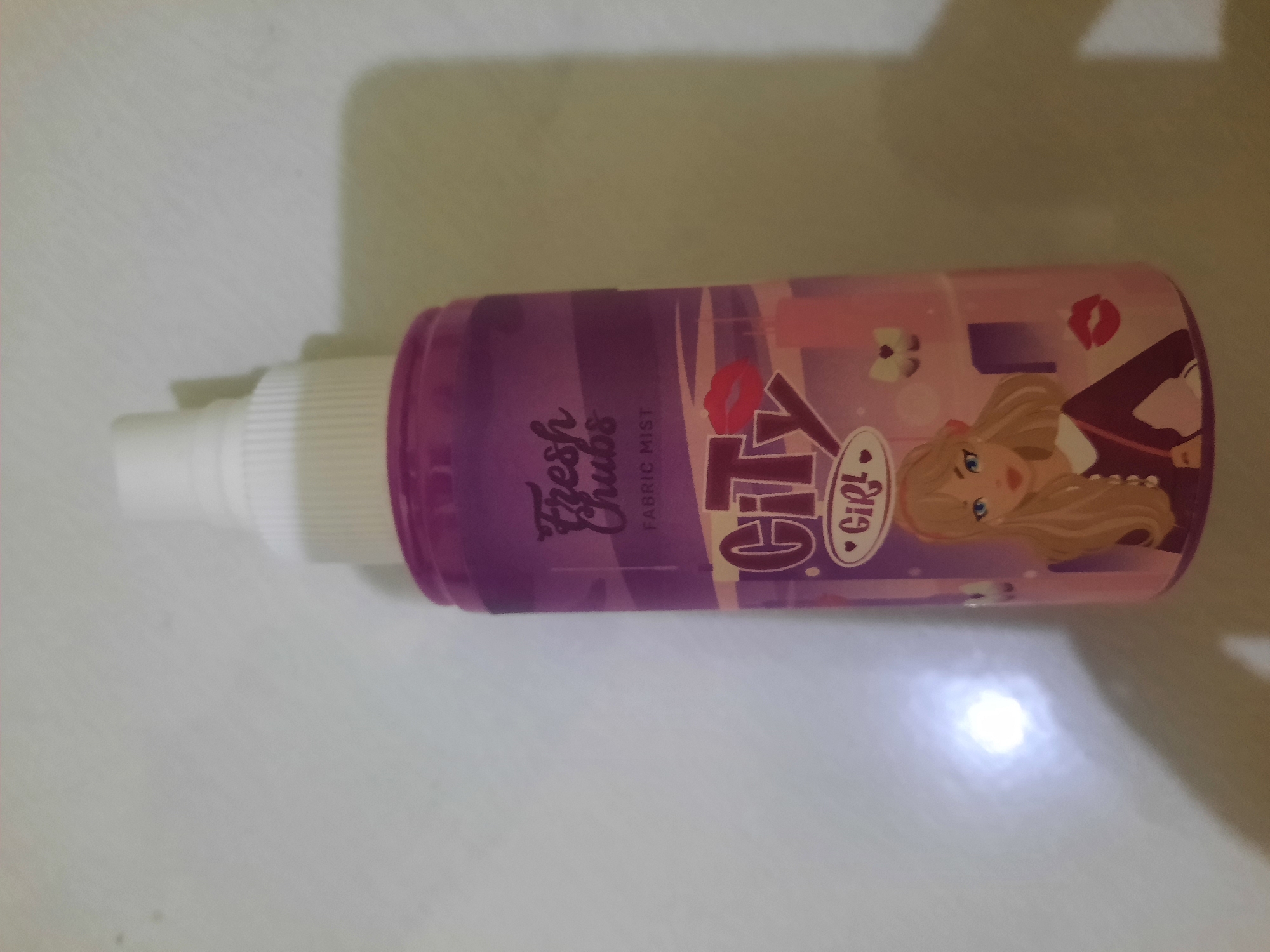A close-up, slightly angled, eye-level shot captures a purple spray bottle with a white cap against a plain, off-white background. The bottle features a colorful label with various graphics and text. At the top of the label, "Fresh Chubs" is written in a stylized font, followed by "FABRIC MIST" below it. A prominent graphic of "City Girl" is displayed in the center, with a stylized red kiss mark above the "i" and a pink heart enclosing "Girl" below the "y". A cartoon illustration of a blonde girl with blue eyes, wearing a dark outfit with a white collar and a red purse, is depicted on the lower portion of the bottle. The background is simple, with subtle shadows cast to the right of the bottle, suggesting an indoor setting. There is a bright, circular glare on the left side of the background, likely a reflection from a light source. There are no discernible elements in the image that would indicate the location in the city of Lhokseumawe, Indonesia.