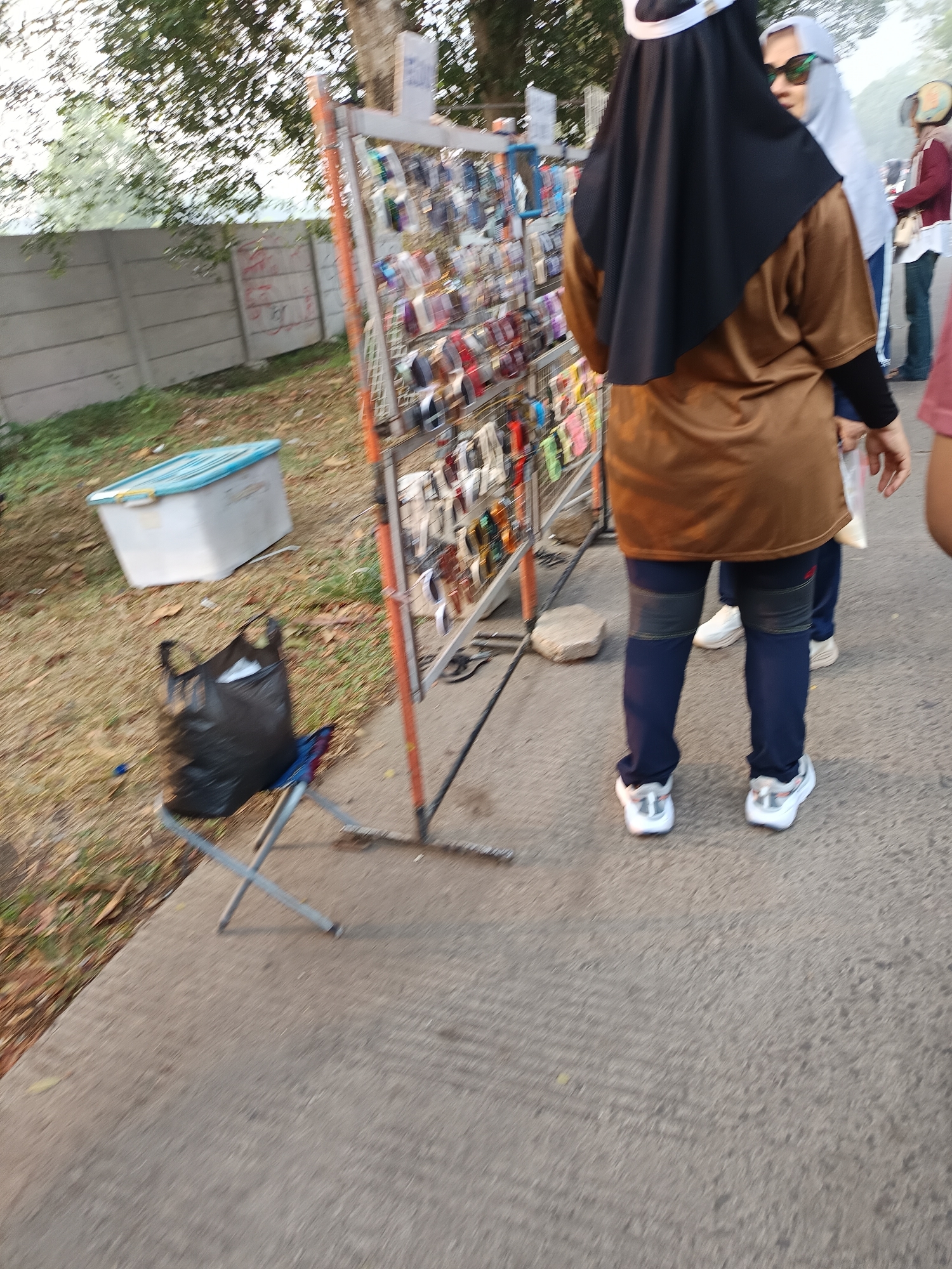 A full shot captured from a low angle shows a woman wearing a brown athletic top, navy blue leggings, and white sneakers, standing in front of a metal display rack filled with various items, possibly sunglasses or phone accessories. She is facing away from the camera, with her back to the viewer, and appears to be browsing the merchandise. To her left, a black plastic bag is placed on the ground next to a small folding chair. In the background, a light-colored storage bin with a blue lid sits on the grassy ground. A concrete wall with graffiti marks is visible behind the bin, and trees and foliage frame the scene. The pavement in the foreground is grey. The overall setting suggests an outdoor market or street vendor area in Sodong, Indonesia, characterized by a casual and bustling atmosphere.