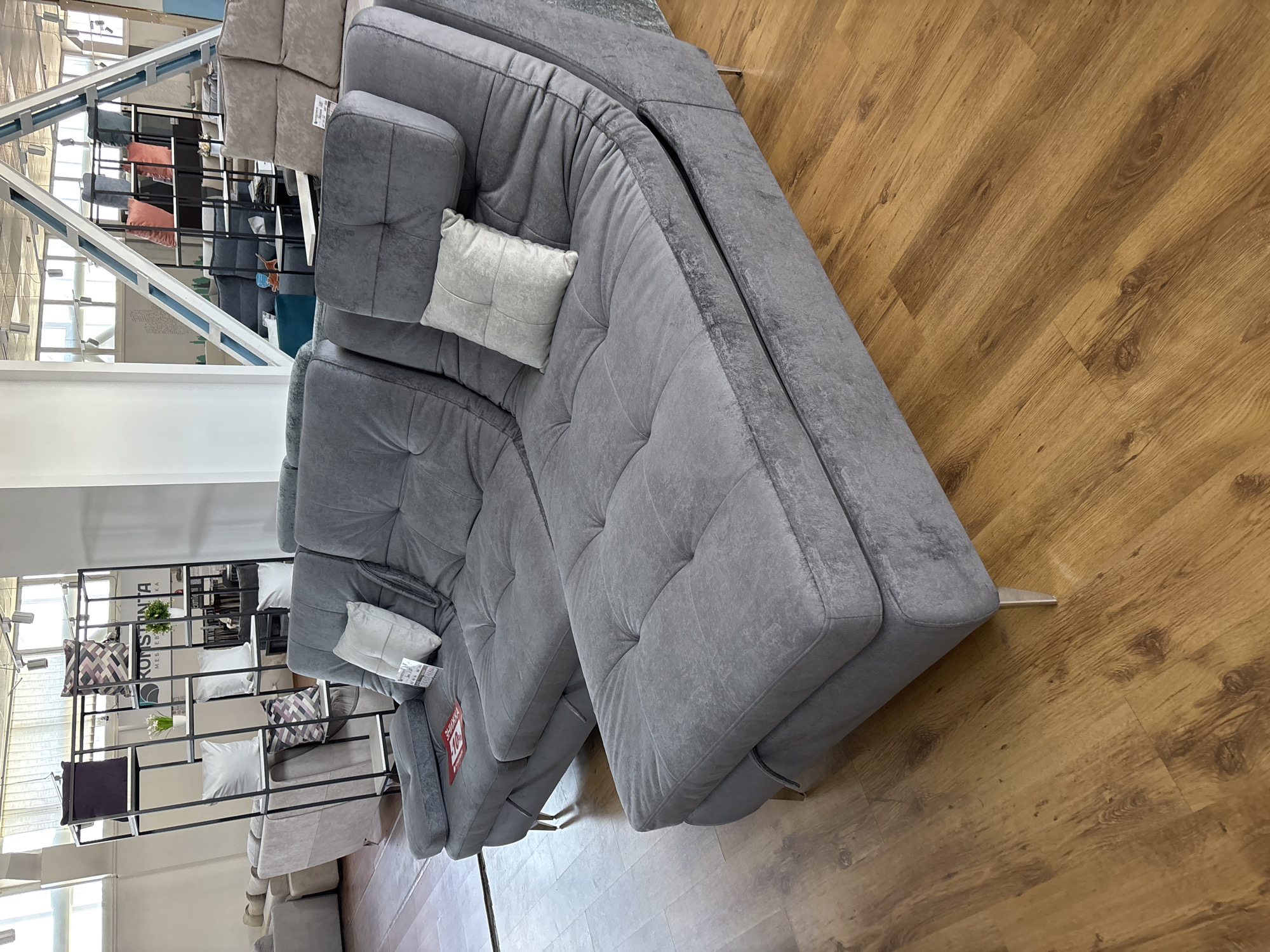 The image depicts the interior of a furniture store located in Zaporizhzhia, Ukraine, showcasing a large, modular gray sectional sofa. The sofa is upholstered in a textured fabric and features multiple cushions, including two decorative pillows. One pillow is light gray and square-shaped with a quilted pattern, while another is off-white and rectangular. A red sale tag is visible on the left armrest of the sofa.
The sofa is positioned on a light brown wooden floor. In the background, several other furniture pieces and display shelves are visible. To the left, a tall, black metal shelving unit holds various decorative items, including pillows with geometric patterns and small potted plants. Further in the background, more sofas and seating arrangements are present, along with large windows that allow natural light to illuminate the space. The overall environment suggests a retail setting with a focus on home furnishings.
