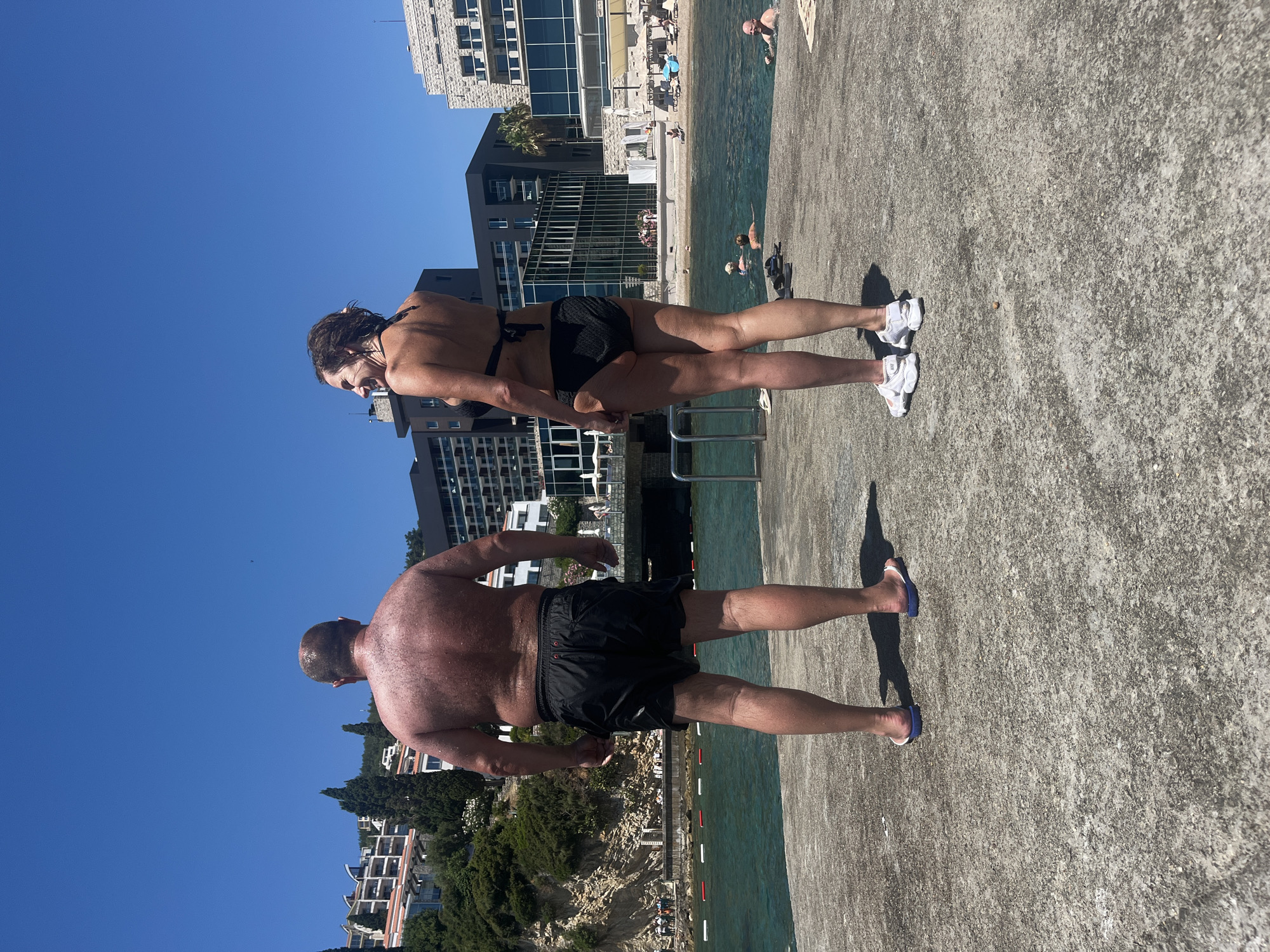 In Budva, Montenegro, two individuals, a man and a woman, stand on a concrete platform overlooking a body of water. The man, viewed from behind, wears black swim shorts and flip-flops. His skin appears tanned and glistening with water. The woman, also viewed from behind, is in a black bikini and athletic shoes. She is looking to her left and smiling. The background features a shoreline with people relaxing on lounge chairs under umbrellas, several buildings, and lush green vegetation on a hillside. The water is a clear turquoise, with buoys marking a swimming area. The sky is a vibrant, cloudless blue.