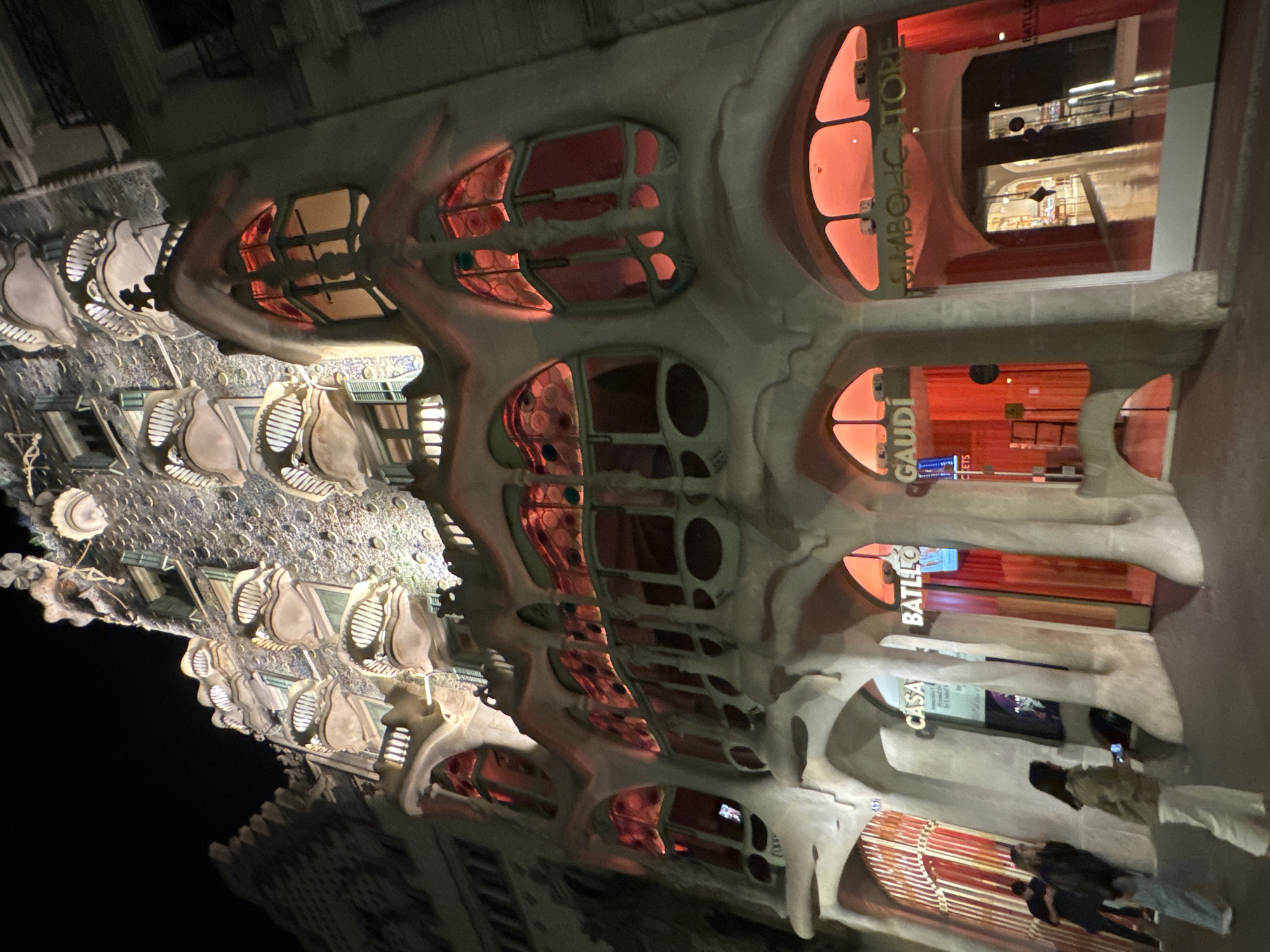 The image shows the facade of Casa Batlló, a renowned building designed by Antoni Gaudí, located on Passeig de Gràcia in Barcelona, Spain. The facade features undulating lines and organic shapes, with bone-like stone details and colorful mosaics. Several windows are visible, some displaying the words "GAUDÍ", "CASA BATLLÓ", and "SIMBOLC STORE." People are visible in the lower right corner, and reflections can be seen in the windows. The image appears to be taken at night, with illuminated windows providing a warm red glow.