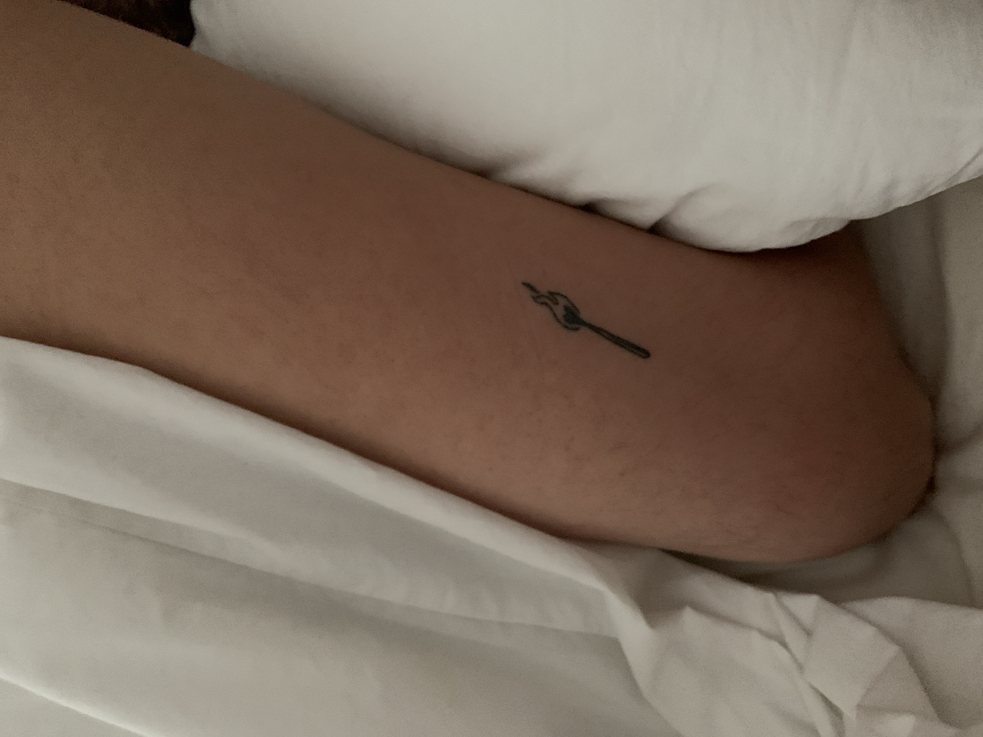An arm with a tattoo of a burning match rests against a white pillow and white bedding. The location cannot be determined from the image.