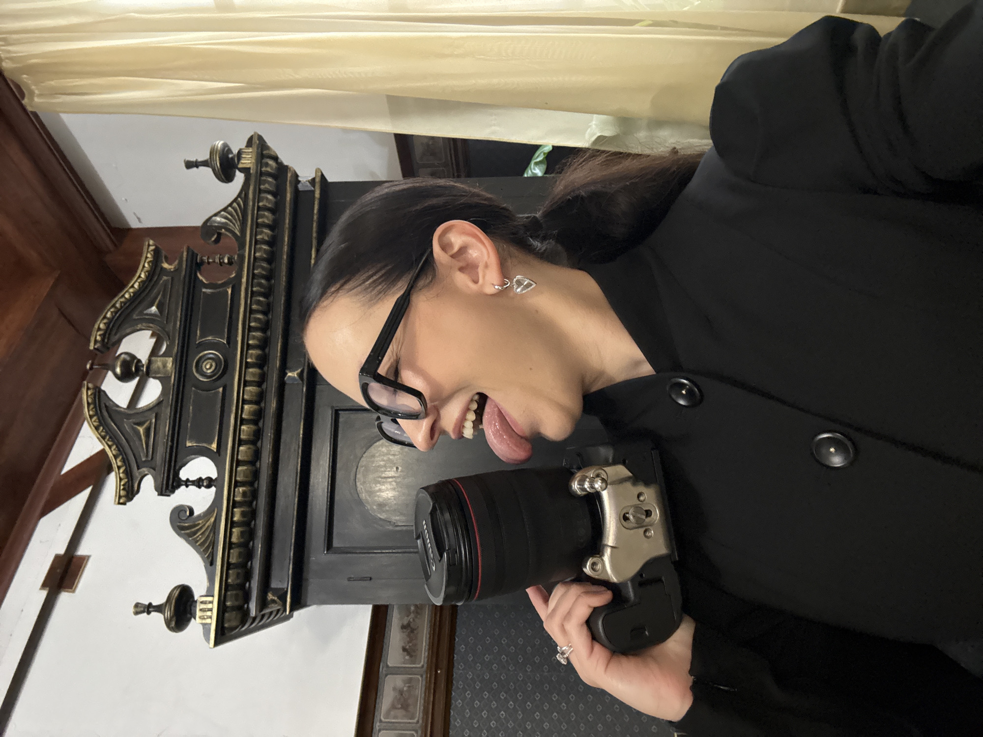 The image shows a woman holding a camera to her face, with her tongue sticking out and her mouth open. She is wearing a black jacket and large black-rimmed glasses. She has dark hair pulled back in a ponytail and a heart-shaped earring. Behind her is a large, ornate dark cabinet with gold trim and what appears to be a ceiling with wooden beams. A light-colored curtain is visible to the right of the cabinet. The overall setting appears to be indoors, possibly in a historical building or museum, given the cabinet's elaborate design. The city of Zavod Mikhailovskii, Russia, is not identifiable from the visual information presented in the image.