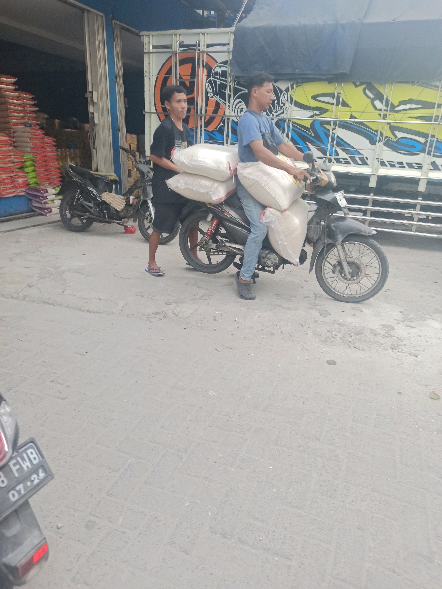 Two young men are on a motorcycle, carrying large sacks of what appears to be rice, in Telagamurni, Indonesia. The man in front is driving the motorcycle, and the man behind him is holding onto the sacks. They are in front of a store with stacks of rice bags outside, and a large truck with graphics is parked behind them. The ground is paved with concrete. The overall scene depicts a commercial area, likely a marketplace or a distribution point for agricultural goods.