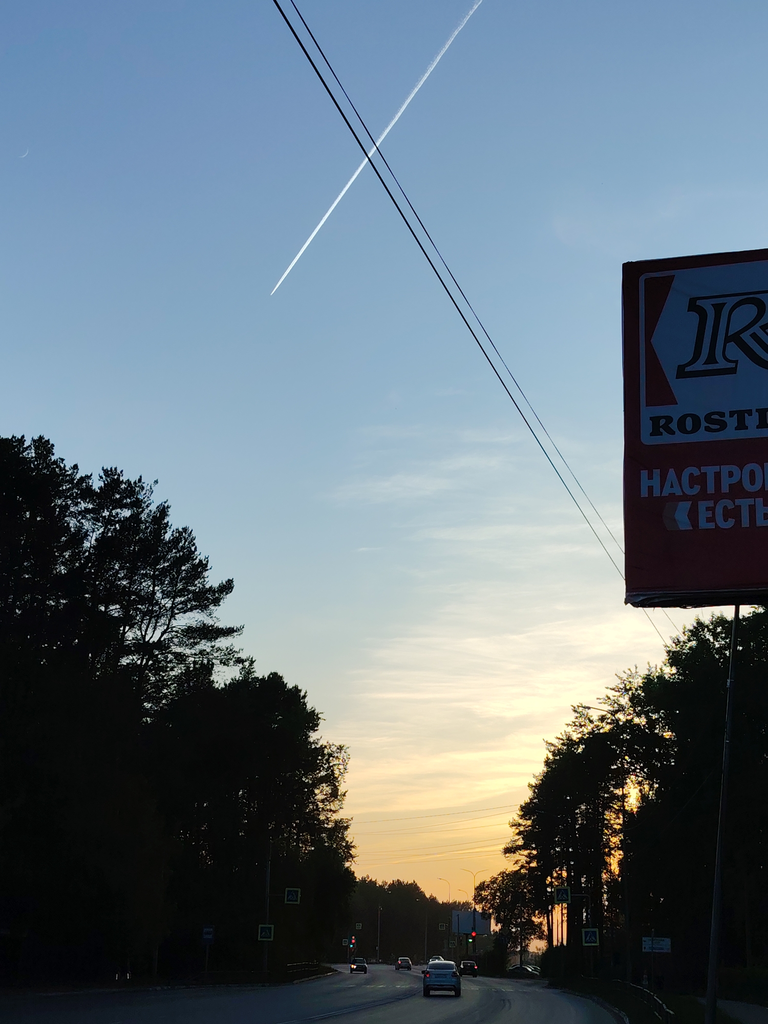 The image shows a view from a road in the city of Chaikovsky, Russia, at sunset or dawn. A crescent moon is visible in the upper left of the blue and light yellow sky, along with the contrails of two crossing aircraft and two utility wires that stretch from the top of the frame. On the right side of the image, a large sign with the partial word "ROSTI" in Cyrillic letters is visible. The word "НАСТРОЕНО" is printed below "ROSTI." The sign is attached to a post, and behind it are dark trees. The road curves gently into the distance. Several cars are visible on the road, including a light-colored sedan in the foreground and other vehicles further away. Dense, dark trees flank both sides of the road, silhouetted against the bright sky. Yellow pedestrian crossing signs and traffic lights are also visible along the roadside.