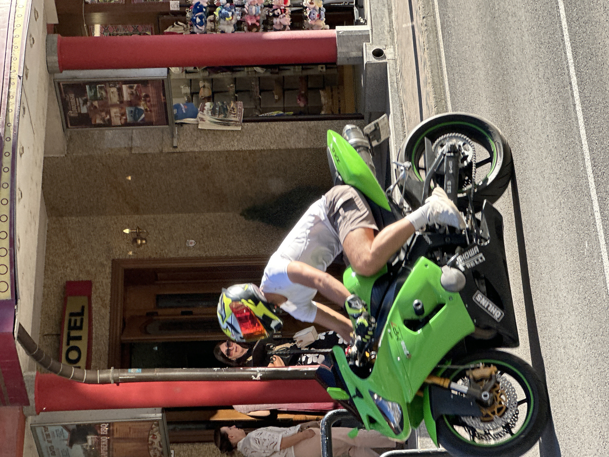 A person wearing a white t-shirt, brown shorts, white socks, and white sneakers is riding a bright green sport motorcycle. They are wearing a yellow and red motorcycle helmet with a mirrored visor. The motorcycle has black accents, a silver exhaust pipe, and the brand "OHLINS" is visible on the side. The rider is in motion on a paved road.
In the background, a building with a textured beige facade has a red pillar. A sign above the entrance reads "OTEL" in yellow letters. Two women are standing near the entrance, one wearing a white patterned top and beige pants, and the other wearing a black dress with a floral pattern and sunglasses. Another building in the background appears to be a shop with shelves displaying various items, including small stuffed animals and mugs. A poster is mounted on the wall. The scene is outdoors, likely on a sunny day, with shadows cast on the ground. The location is the city of Veliko Tarnovo, Bulgaria.
