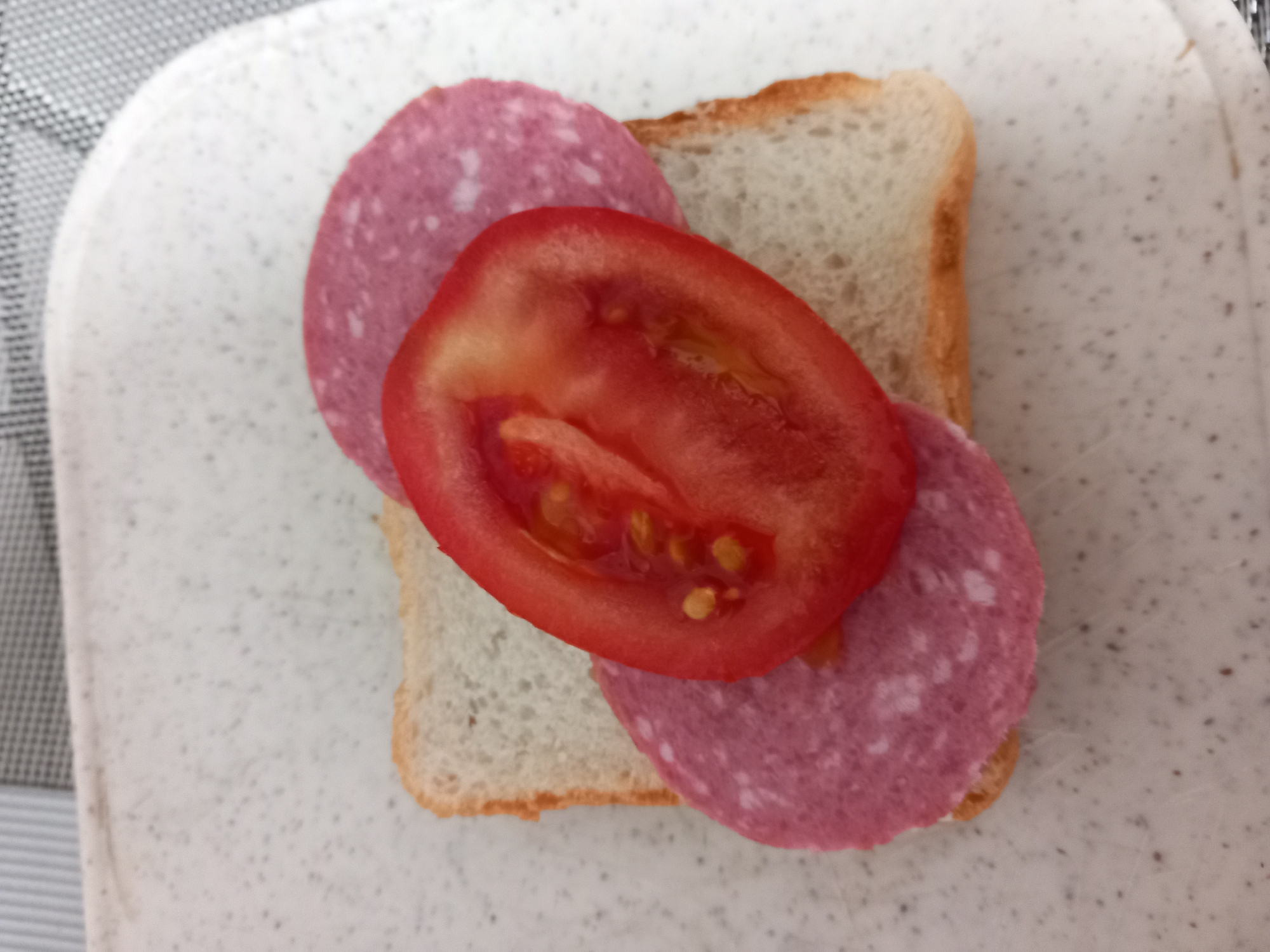 A close-up, overhead shot shows a slice of white bread topped with two slices of salami and one slice of tomato. The salami slices are pink with visible fat marbling. The tomato slice is red and juicy, with seeds visible. The sandwich is on a white speckled cutting board. The background is blurred, with stripes of grey and white fabric.