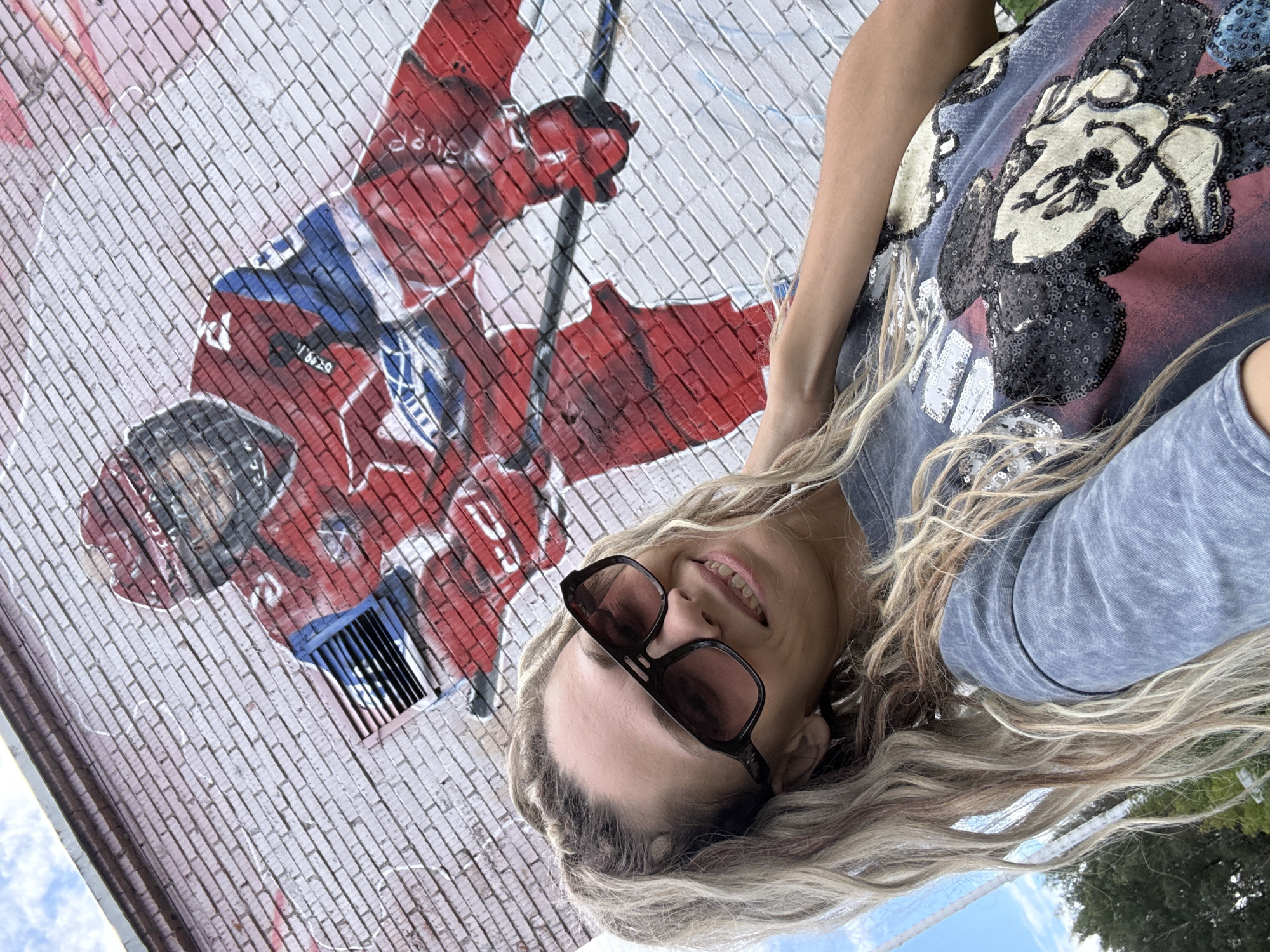 In Rusanovo, Russia, a woman with blonde hair and dark sunglasses takes a selfie in front of a brick wall mural of a hockey player. She is wearing a gray t-shirt with a sequined Mickey Mouse graphic. The mural depicts a player in a red and blue uniform, with a hockey stick. The sky is visible in the upper left corner, with some clouds.