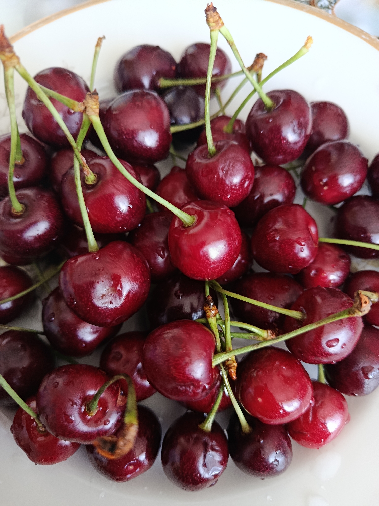 A close-up, high-angle shot captures a pile of dark red cherries with visible water droplets in a white bowl. The cherries are clustered together, with some stems still attached, and their glossy surfaces reflect the light. The bowl itself appears to have a subtle pattern around its rim. There are no people present in the image, and the scene is devoid of any urban or natural landmarks. The image was taken in Svietlahorsk, Belarus.