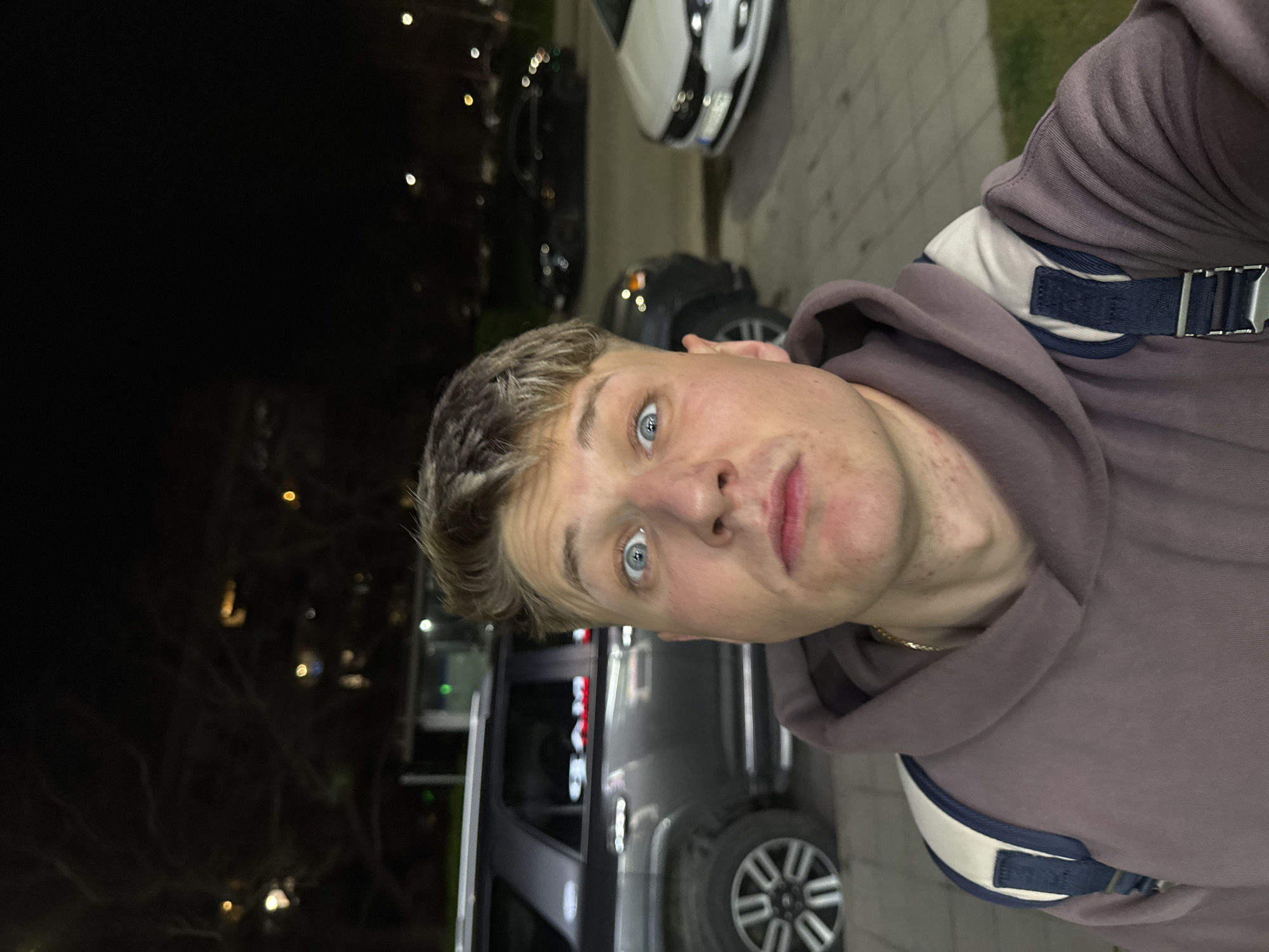 A selfie of a young male with blonde hair and blue eyes, wearing a hooded sweatshirt and a backpack, taken outdoors at night. The background suggests a city street, with cars parked along a curb and streetlights visible in the dark. The location is possibly in Plovdiv, Bulgaria.