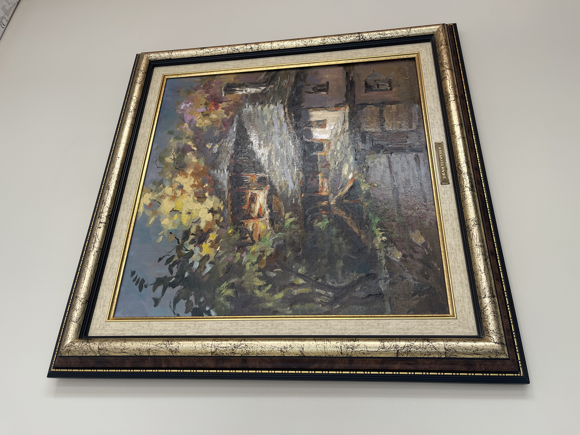 The image shows a framed oil painting hanging on a white wall. The painting depicts a house or building partially obscured by trees and foliage. The color palette is dominated by greens, browns, and yellows, suggesting an autumn or late summer scene. The painting has a signature on the right side, identified as "Ivan Portilla" on a small plate fixed to the frame. The frame is ornate, consisting of multiple layers in gold, silver, brown, and black. The location is not discernible from the image itself.