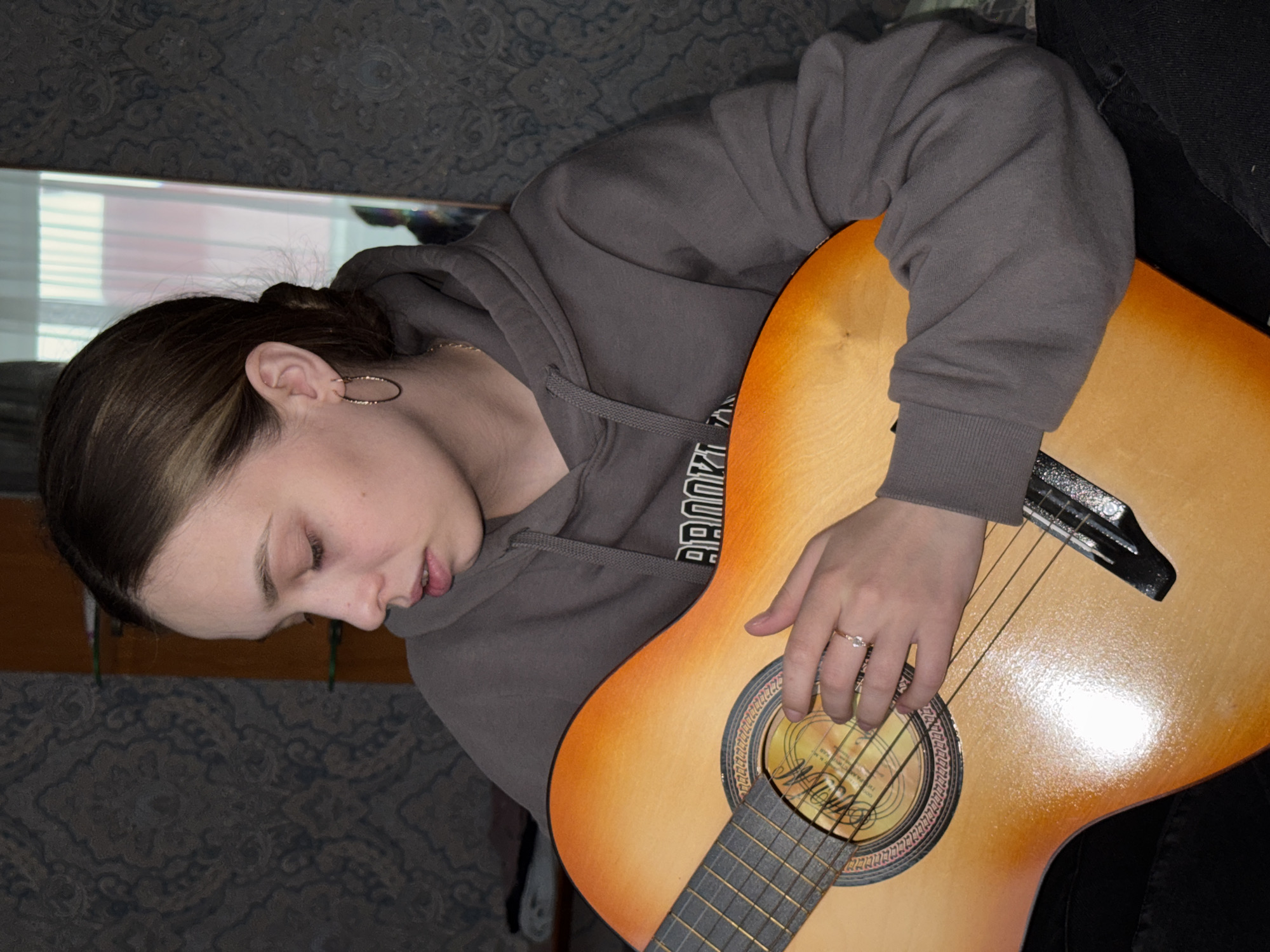 A young woman is playing an acoustic guitar. She has brown hair pulled back, hoop earrings, and is wearing a gray hoodie with the word "BROOKLYN" printed on it. Her left hand is fretting the strings, and she is wearing a ring on her ring finger. The guitar is light orange with a dark fretboard. The background includes a patterned wall and a glimpse of a window and wooden surface, possibly reflecting in a mirror. The location cannot be confirmed as Прикамский, Russia based on the visual information.