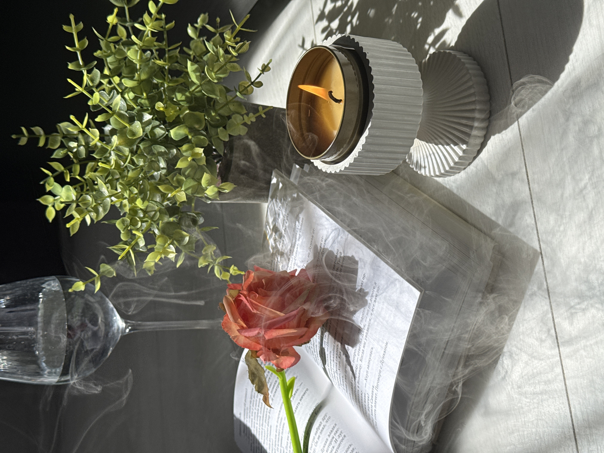 An open book with a single orange rose placed on its pages lies on a light-colored wooden surface. Wisps of smoke or vapor rise from the book and surround the rose, creating an ethereal effect. To the right of the book, a lit candle in a decorative white holder emits a warm glow, casting a shadow on the surface. Behind the book and candle, a glass of water and a potted green plant are visible. Sunlight streams in from the right, creating strong shadows and highlights. The background is dark and out of focus. There are no people present in the scene.