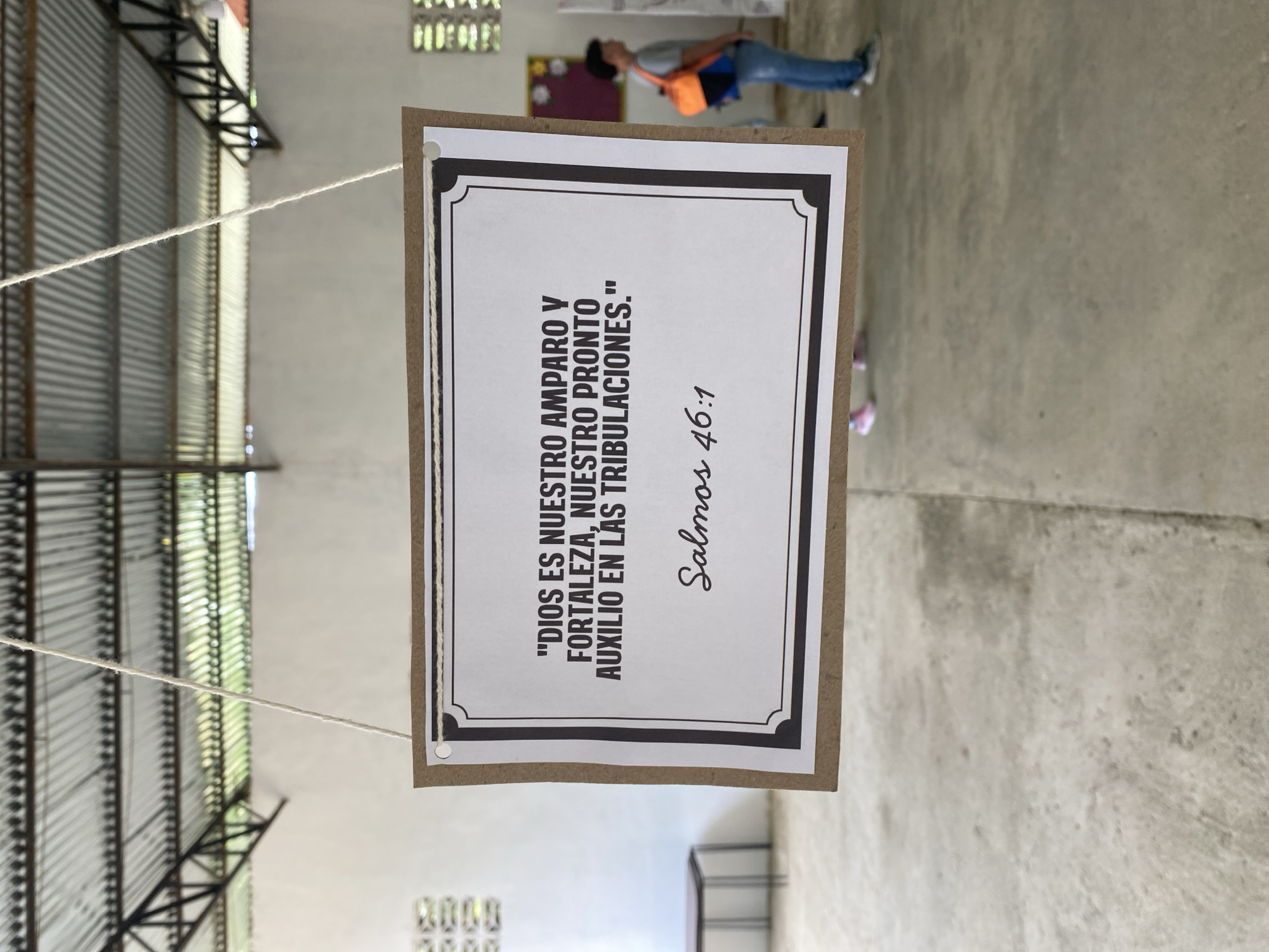 A photograph depicts a sign hanging by strings in what appears to be an indoor setting. The sign is composed of a piece of brown paper as its base, with a white sheet printed with black ink affixed to it. The text on the sign, written in Spanish, reads, "DIOS ES NUESTRO AMPARO Y FORTALEZA, NUESTRO PRONTO AUXILIO EN LAS TRIBULACIONES." followed by "Salmos 46:1." The sign has a thin black decorative border around the white sheet.
In the background, the ceiling has a corrugated metal structure, and there is a person wearing blue jeans, a gray shirt, and an orange bag standing in the distance. The floor appears to be a gray concrete slab.
The image was taken in the city of Tinaquillo, Venezuela.