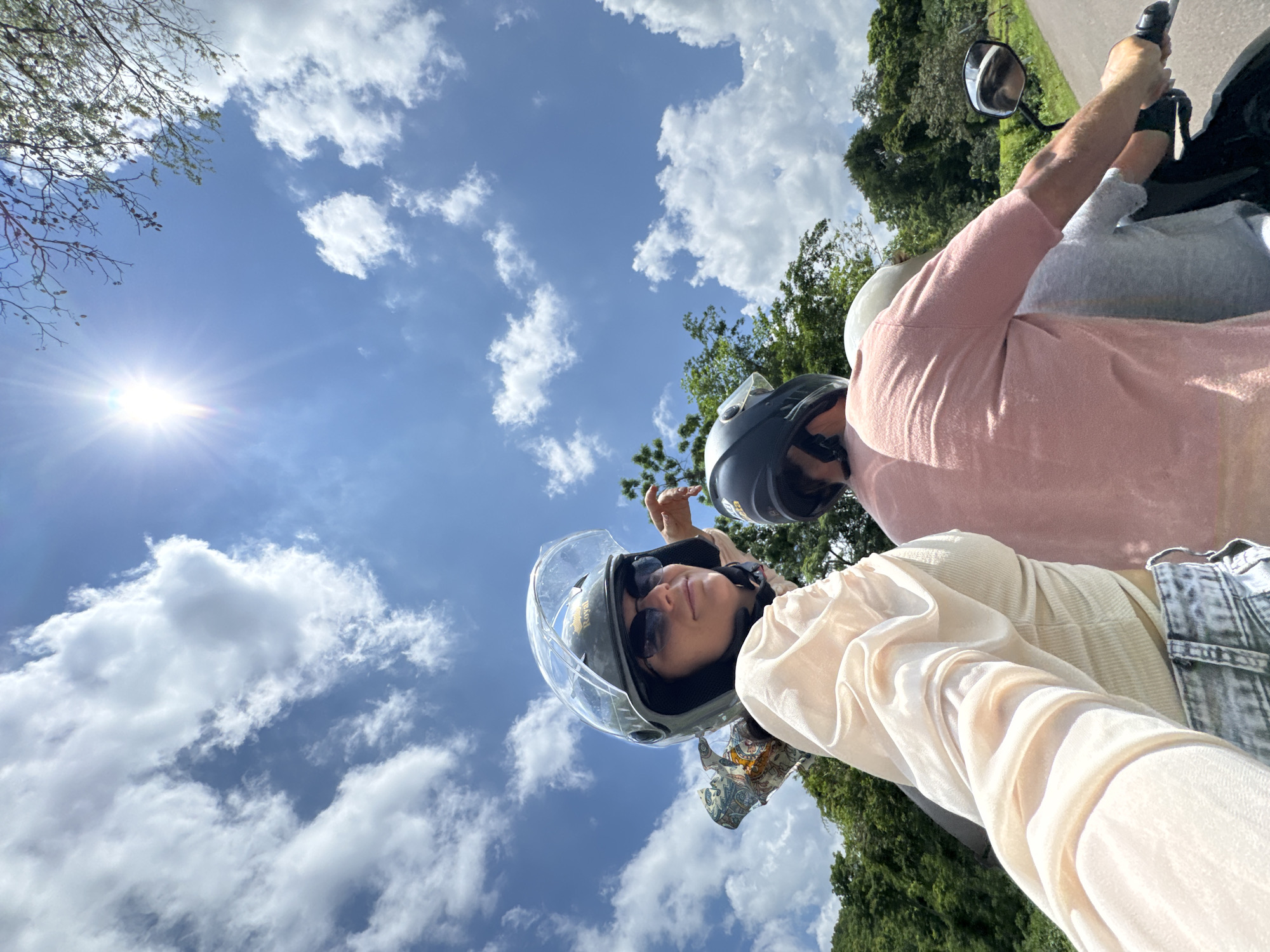The image shows three individuals riding on a motorbike under a bright, sunny sky with scattered clouds. The person in the foreground, wearing a light-colored top and jeans, is looking towards the camera and wearing a helmet and sunglasses. To their right, another person is wearing a pink top and a helmet, and a third person, partially visible, is wearing a light gray shirt and a helmet. They are on a paved road surrounded by lush green trees and vegetation. The overall scene suggests a scenic journey on a motorbike, possibly in a rural or natural area. The specific location in Ham Thuan Bac, Vietnam, is not visually discernible from the image alone but could be inferred from metadata or external context.