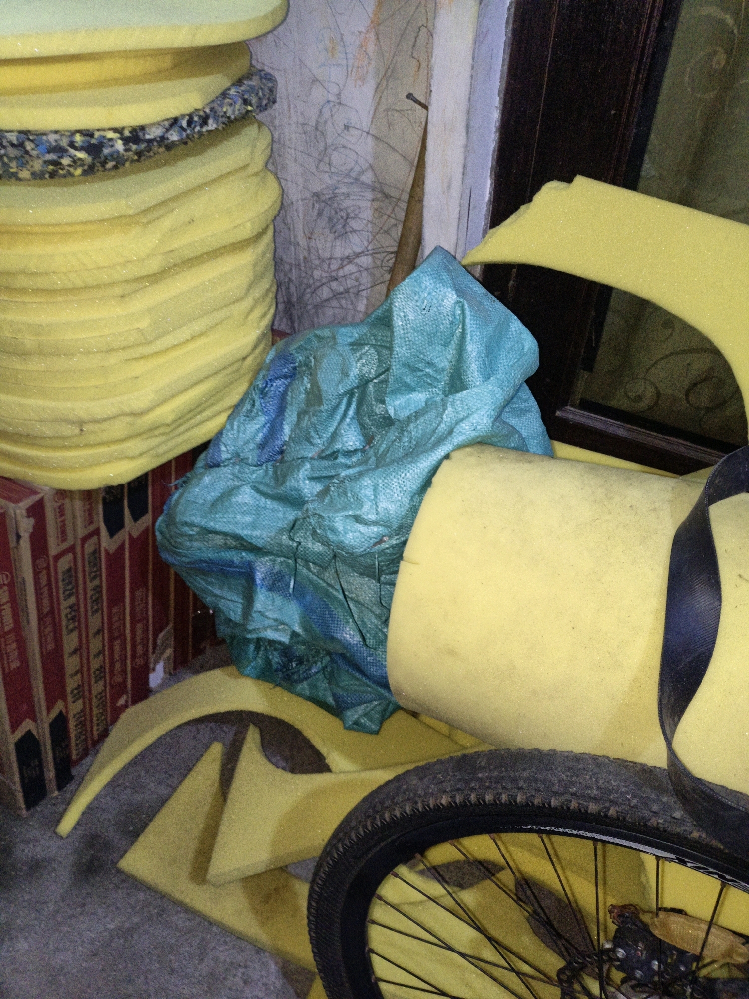 The image shows a pile of yellow foam padding and a crumpled teal tarp. The foam padding is stacked in large, circular pieces and some curved pieces are lying on the floor. The teal tarp is draped over some of the foam. In the foreground, a bicycle wheel with a black tire is visible, resting on some of the foam padding. Behind the foam and tarp, there are cardboard boxes and a dark wooden structure, possibly a door or window frame. The overall environment appears to be an indoor storage or workshop area, with a textured wall in the background. There are no people visible in the image. The scene is lit by artificial light, casting shadows. The location is Cikal, Indonesia.