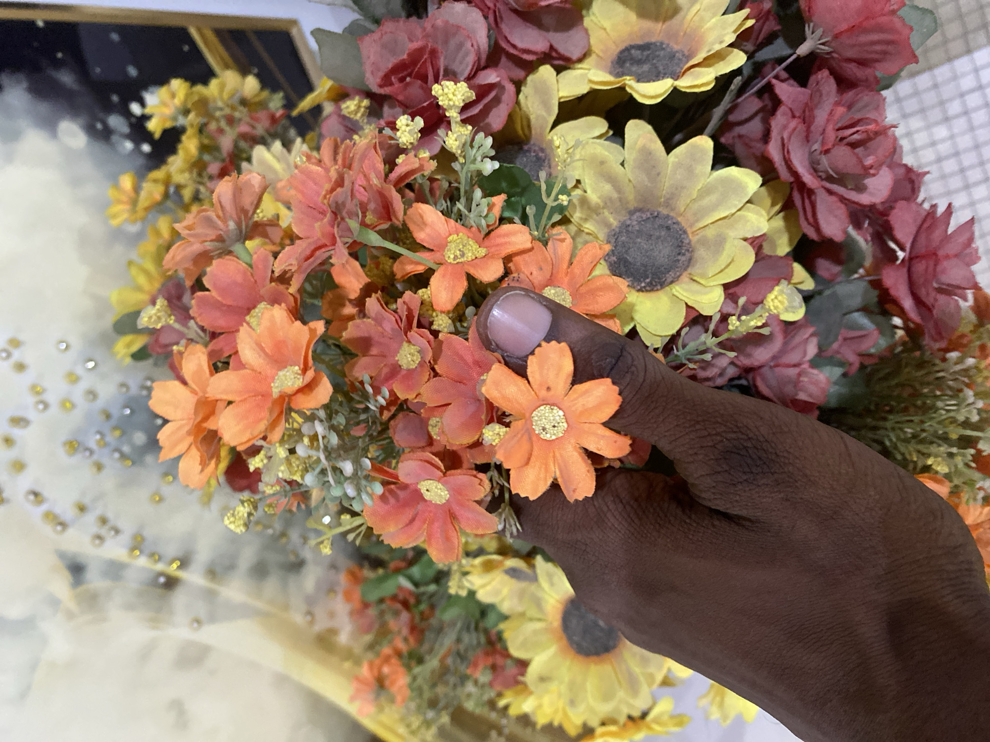 A hand with dark skin and a light-colored fingernail is holding a bouquet of artificial flowers. The bouquet contains flowers of various colors, including orange, yellow, and burgundy. Some of the flowers are small and daisy-like, while others are larger and resemble sunflowers or roses. The background of the image is out of focus and appears to be a wall with some decorative elements. The image was taken in Giginyu, Nigeria.
