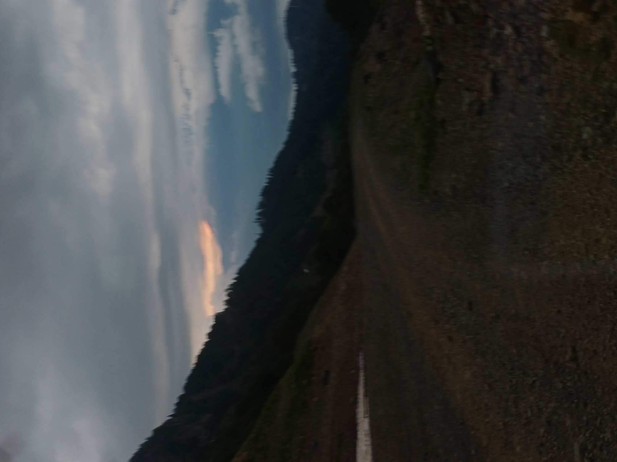The image shows a dark, blurry landscape, likely taken at dusk or dawn. The foreground is a rough, gravelly surface, possibly a road or trail, with some patches of lighter colored material that could be snow or dirt. The middle ground features a dark, tree-covered hillside sloping upwards, with a lighter patch visible on the left side. In the background, dark mountains are silhouetted against a cloudy sky. The sky itself is overcast with dark grey clouds, but there are hints of warmer light breaking through in the distance, suggesting a sunrise or sunset. The overall impression is one of a natural, remote environment. The location is specified as Estosadok, Russia, a mountain resort area.