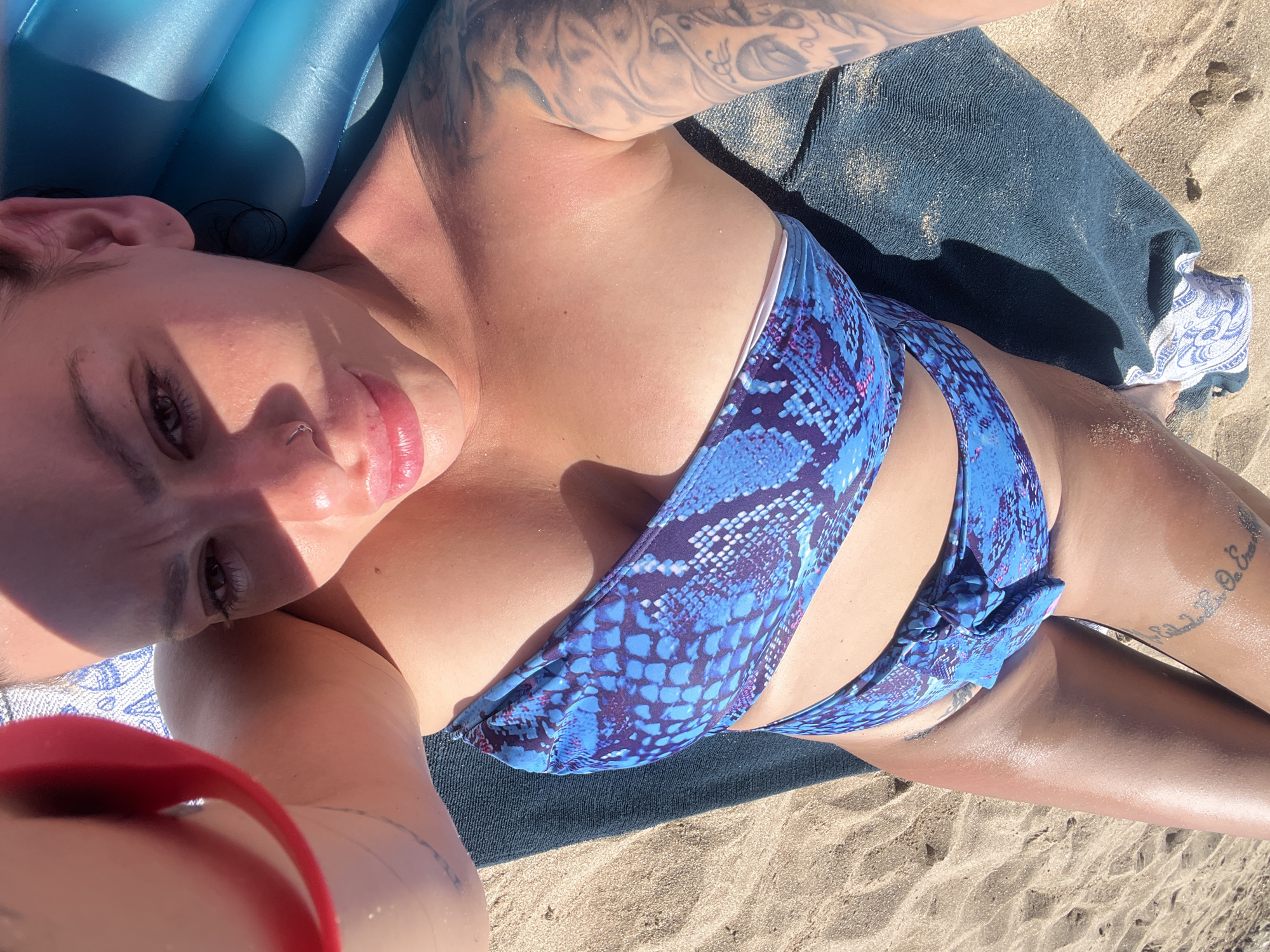 The image shows a person lying on a beach, viewed from above. The person is wearing a blue and purple patterned bikini. Their face is partially visible, with eyes closed and lips slightly parted. Sunlight casts shadows across their face and body, indicating it is daytime. Tattoos are visible on their arm and leg. A blue inflatable mat and a dark towel are present, along with sandy ground. The scene suggests a leisurely day at the beach in Sozopol, Bulgaria.