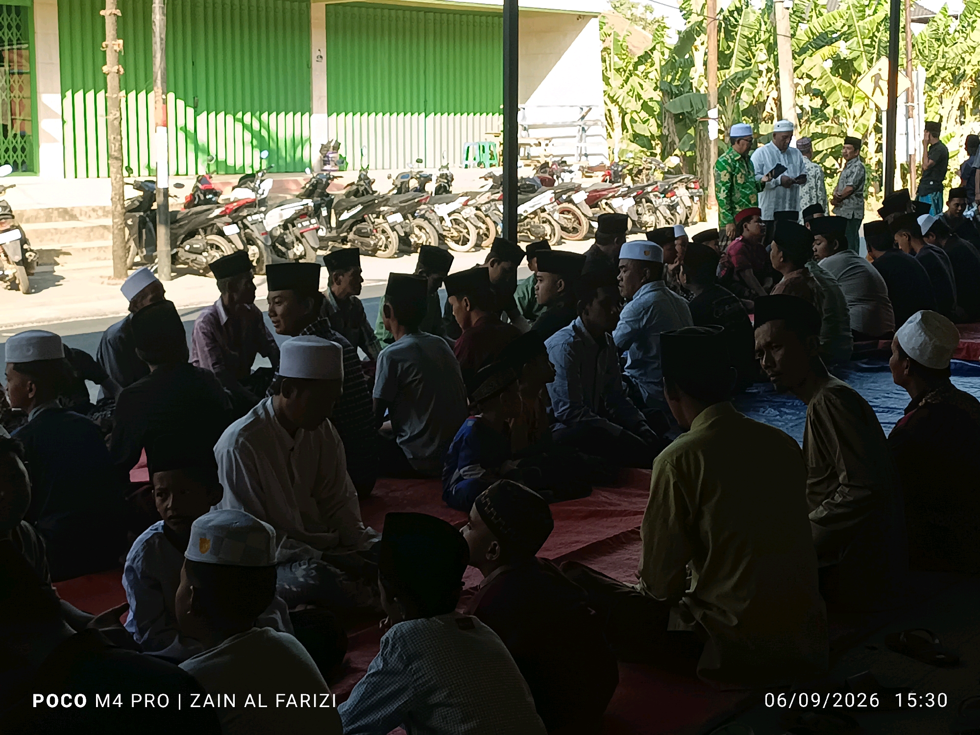 In the city of Cendagah, Indonesia, a group of people are gathered outdoors, with a focus on a man in the foreground wearing a white cap. The scene appears to be a community event or gathering, possibly religious in nature, given the attire of some individuals. In the background, motorcycles are parked in rows, suggesting transportation for attendees. Behind the motorcycles, a building with a green corrugated metal facade is visible, with a white awning or roof structure extending outwards. The ground is covered in red mats, indicating a designated seating area. The lighting suggests it is daytime, with shadows indicating the sun's position. Several individuals are seen wearing peci hats, common headwear in Indonesia. The overall impression is one of a communal event taking place in an open, outdoor space.