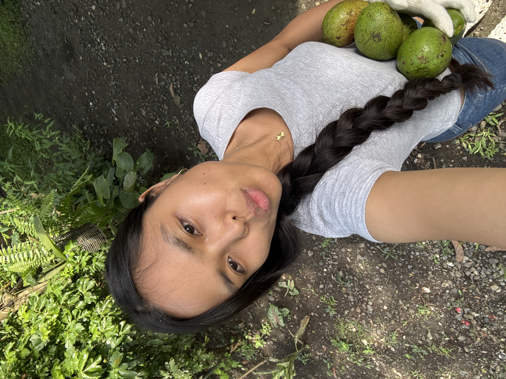 The image shows a woman outdoors with green avocados. She is wearing a gray t-shirt, blue jeans, and a gold necklace, with her dark hair braided. She is holding the avocados on or near her lap. The ground is a mixture of dirt, gravel, and vegetation, including ferns and other green plants. The location is suggested to be Carmen, Philippines, based on available information, though this is not directly verifiable from the image.