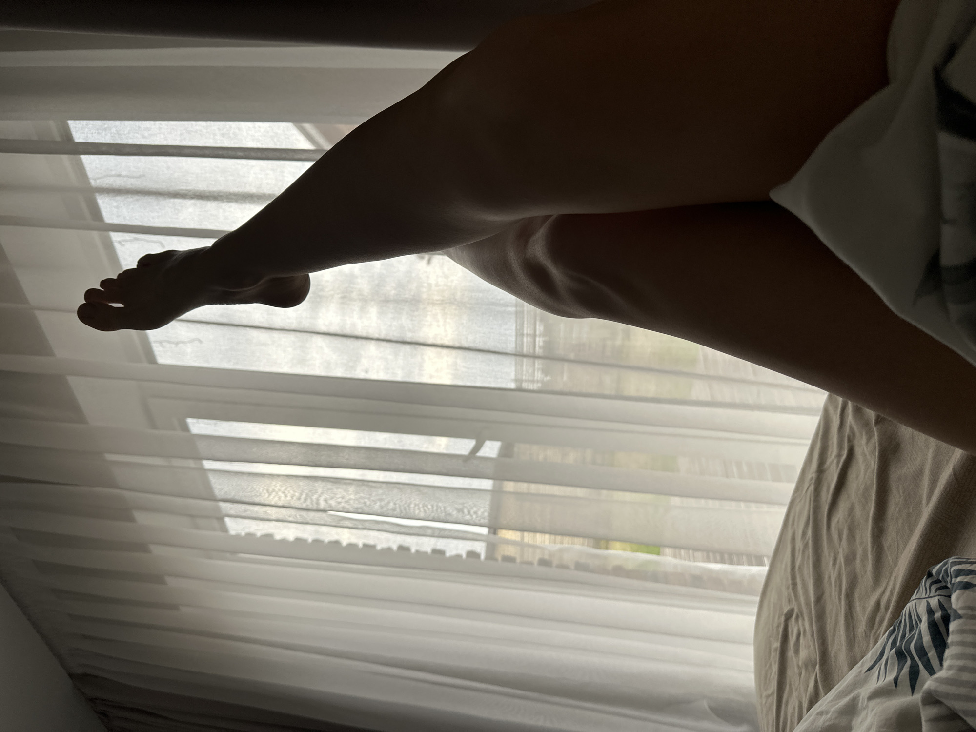 The image shows a person's legs and foot near a window covered with white blinds and a sheer curtain. The legs are bent, with one leg crossed over the other. The foot is visible near the window. Part of a bed with beige and blue-striped bedding is also visible. The location is not discernable from the image, therefore it is impossible to confirm the location is in the city of Chaikovsky, Russia.