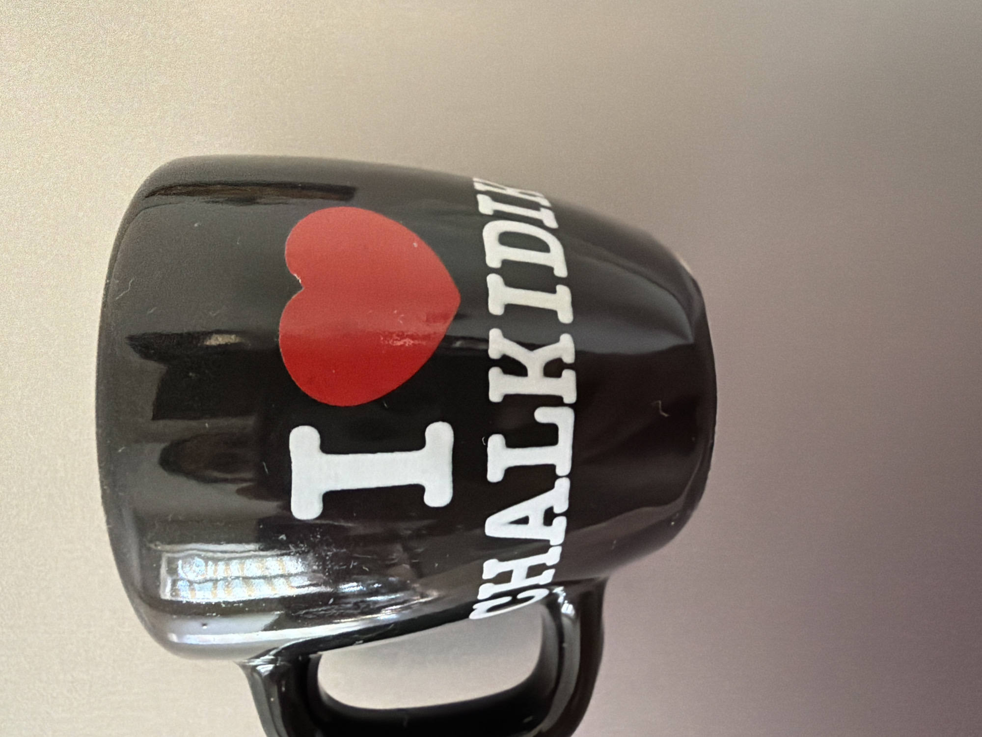 A black mug with white lettering and a red heart is placed on a light-colored surface. The lettering on the mug reads "I CHALKIDI". The mug has a handle on the left side. The background is blurred and out of focus. There is no indication of location in Pazardzhik, Bulgaria.
