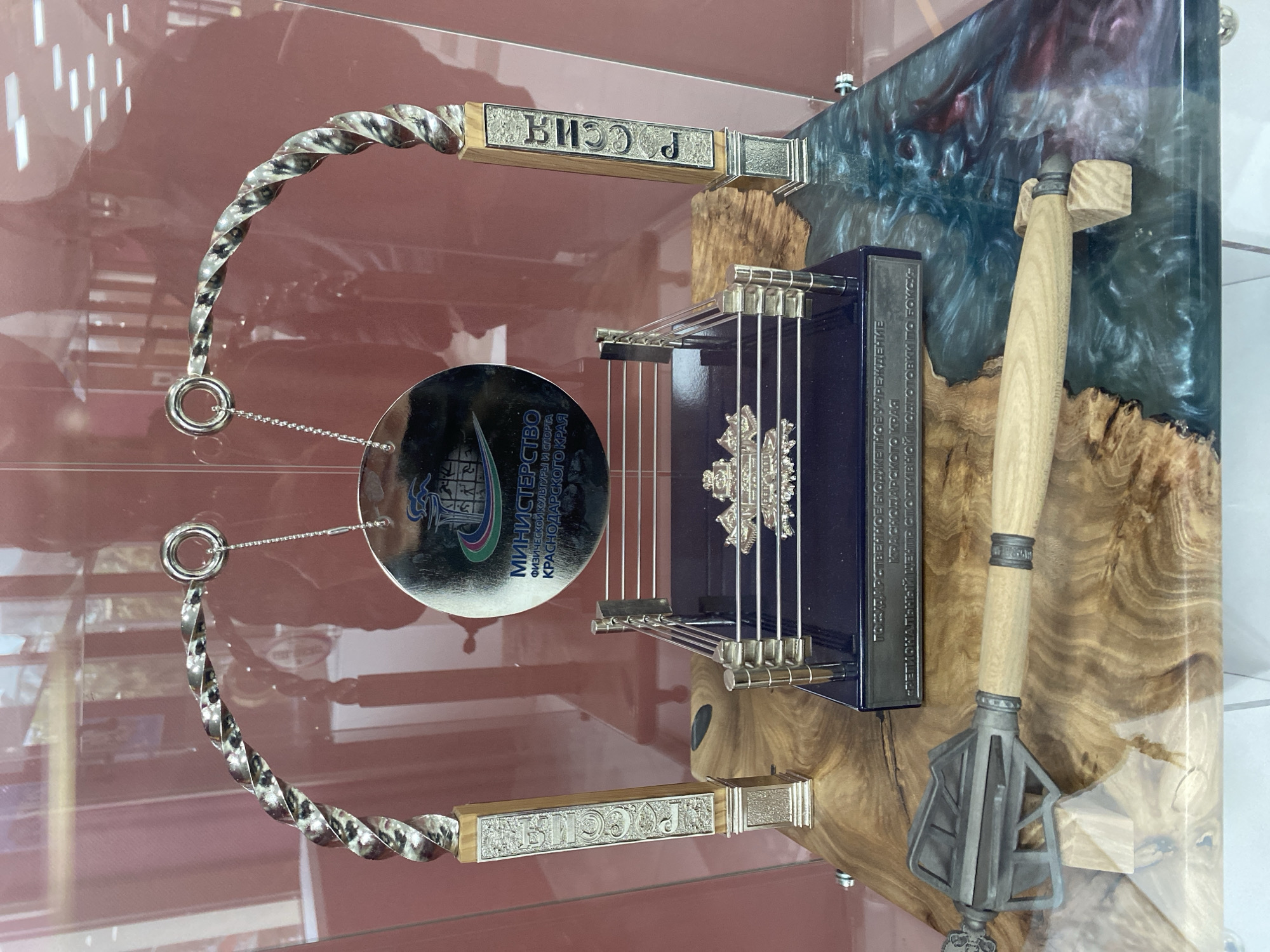 This image depicts a decorative display, likely housed within a museum or display case, as suggested by the lighting and glass reflections. It features a variety of ornate objects arranged on a base that combines light wood with resin. The artifacts include:
1. **A balalaika-like instrument**: A blue rectangular box-shaped musical instrument that resembles a balalaika is centrally placed. It features silver-colored strings and ornate decorations.
2. **Decorative blades**: Attached to the sides of the instrument, there are two twisted silver-colored blades with wooden handles. "PO.CO" is inscribed on one handle and "РОССИЯ" (Russia) is on the other.
3. **A reflective disk**: A circular reflective disk is hanging by chains from the top blade and features an emblem and text in Russian. It reads "МИНИСТЕРСТВО ФИЗИЧЕСКОЙ КУЛЬТУРЫ И СПОРТА КРАСНОДАРСКОГО КРАЯ", translating to the Ministry of Physical Culture and Sports of the Krasnodar Region.
4. **A mace**: To the right, there's a wooden mace with a decorative silver-colored head resting against the resin background.
The presence of Russian text, specifically referencing the Krasnodar Region, suggests that the objects displayed are associated with Russian culture, history, or government. The display likely represents cultural or historical artifacts from the region, possibly related to sports or physical culture. The display's location is possibly within the city of Sochi, Russia, due to the inclusion of text on the display about sports and physical culture, which are common associations with the city of Sochi in Russia.