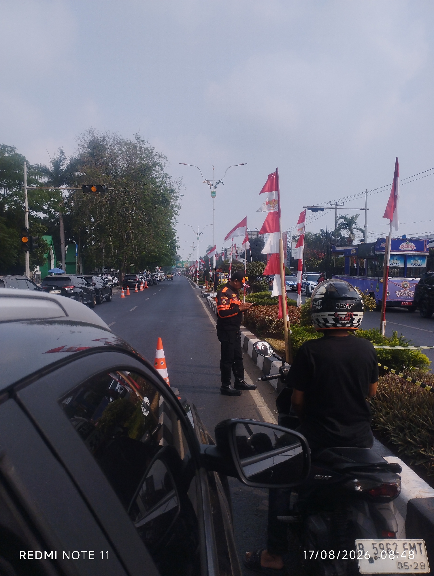 The image depicts a street scene in Karawang, Indonesia, under a hazy sky. In the foreground, the rear of a black car is visible, reflecting the surroundings. A man wearing a black t-shirt and helmet sits on a scooter to the right, facing away from the camera. Further down the road, a person in an orange and black uniform, possibly a traffic officer or event staff, stands holding a mobile phone and looking towards the ground. Several Indonesian flags are displayed along the roadside. Cars are lined up on the road, suggesting a traffic jam or a planned event. Traffic cones are placed along the edge of the road. In the background, trees, streetlights, and buildings can be seen. A timestamp "17/08/2026 08:48" and license plate "B 5962 EMT 05-28" are visible on the scooter. The text "REDMI NOTE 11" is also present in the lower left corner.