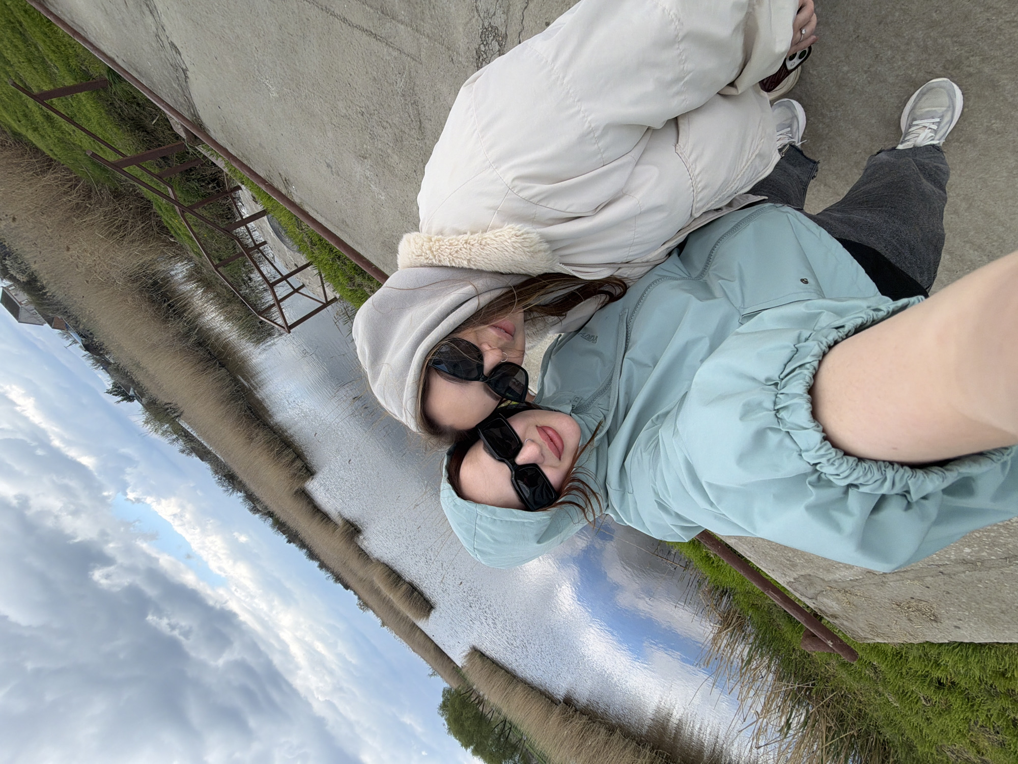 Two individuals are posing for a selfie. One is wearing a white jacket with a fur-lined hood and dark sunglasses. The other is wearing a light blue hooded jacket and dark sunglasses. They are standing on a concrete surface next to a body of water with tall reeds growing along the bank. The sky is cloudy. A rusty metal structure is visible near the edge of the concrete. The location is identified as Unknown, Unknown.