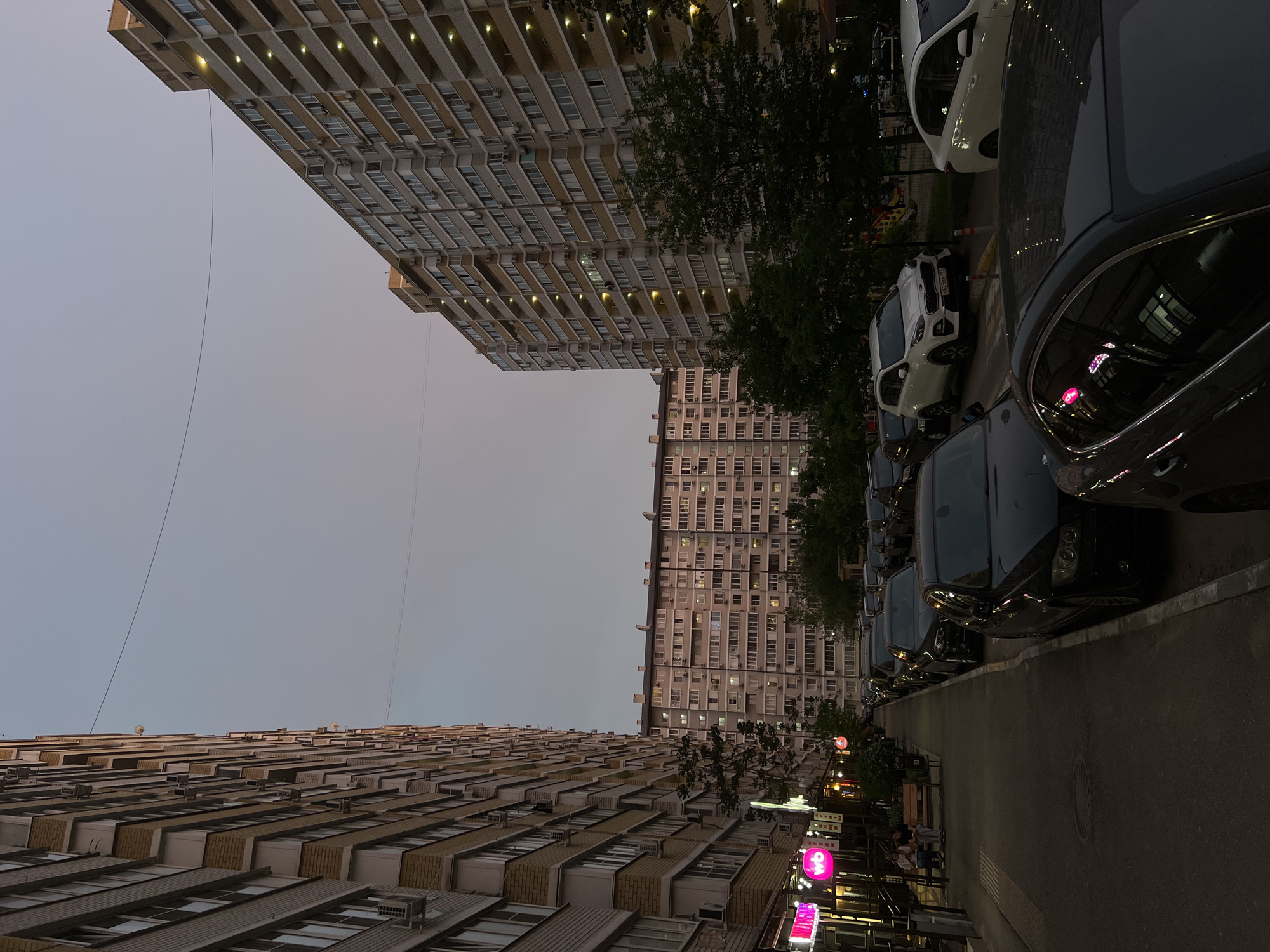 The image depicts a street scene in Krasnodar, Russia, at dusk. Two large, multi-story apartment buildings dominate the view, rising vertically on opposite sides of the frame. The buildings are characterized by rows of balconies and windows.
Parked cars line the right side of the street, their dark surfaces reflecting the ambient light. A sidewalk runs along the right edge of the frame, with shops and businesses visible behind it. Neon signs, including a pink sign, illuminate the storefronts. The sky is a gradient of muted colors, suggesting either sunrise or sunset.