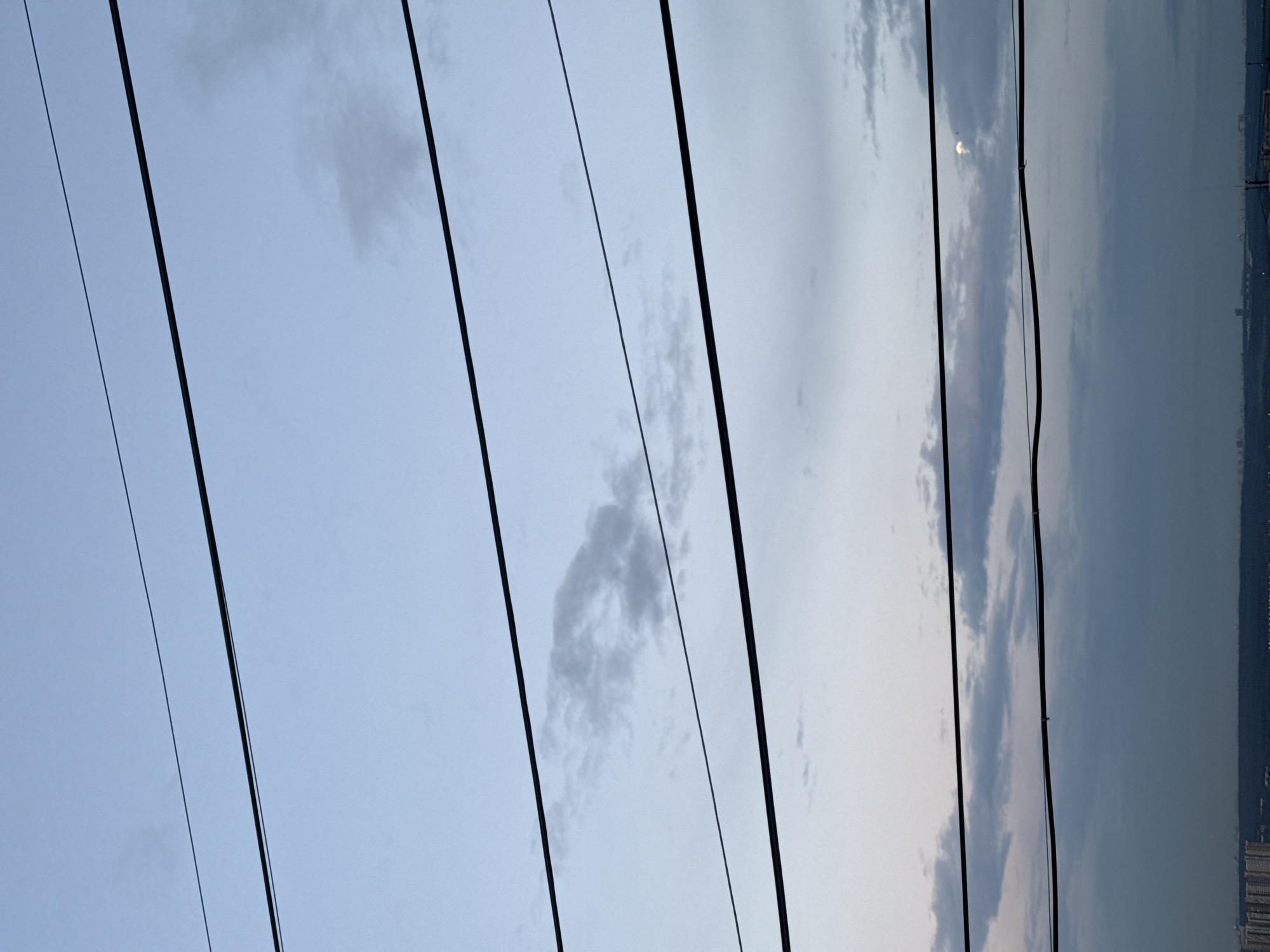 The image depicts a sky with scattered clouds and power lines stretching across the frame. The sun is partially visible through the clouds, suggesting it is either sunrise or sunset. The sky transitions from a light blue on the left to a darker, hazy blue on the right, where a distant city skyline is visible. The power lines are dark and angled, creating a linear pattern against the sky. The overall mood is serene and somewhat vast, with the focus on the expanse of the sky and the man-made elements that punctuate it. No people are visible in the image. The location is Otradnoye, Russia, which could explain the urban skyline in the distance. The time of day is likely early morning or late evening, given the lighting conditions and the presence of the sun partially obscured by clouds. The weather appears to be partly cloudy. There is no visible text in the image.