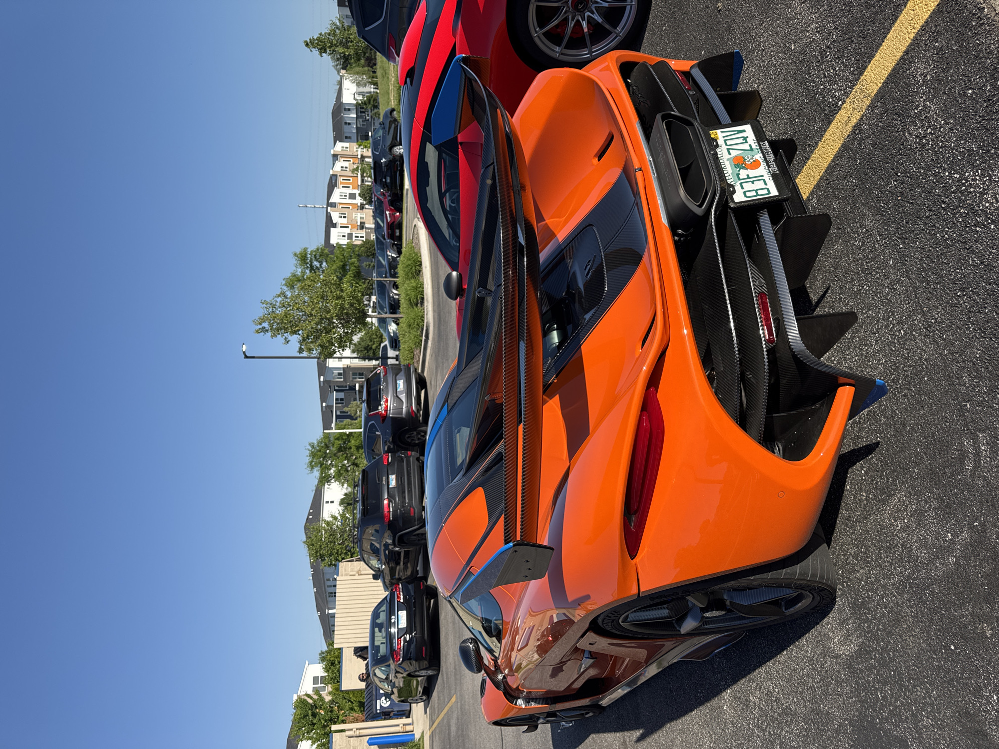 The image shows the rear end of an orange sports car with a large carbon fiber wing, parked in a parking lot. The car has a Florida license plate that reads "BFZE28." A red car is partially visible behind the orange car, as are several other cars of different colors parked in the distance. The background includes a row of houses and a clear blue sky. The parking lot has yellow lines dividing parking spaces. This photograph was taken in Warrenville, United States of America.