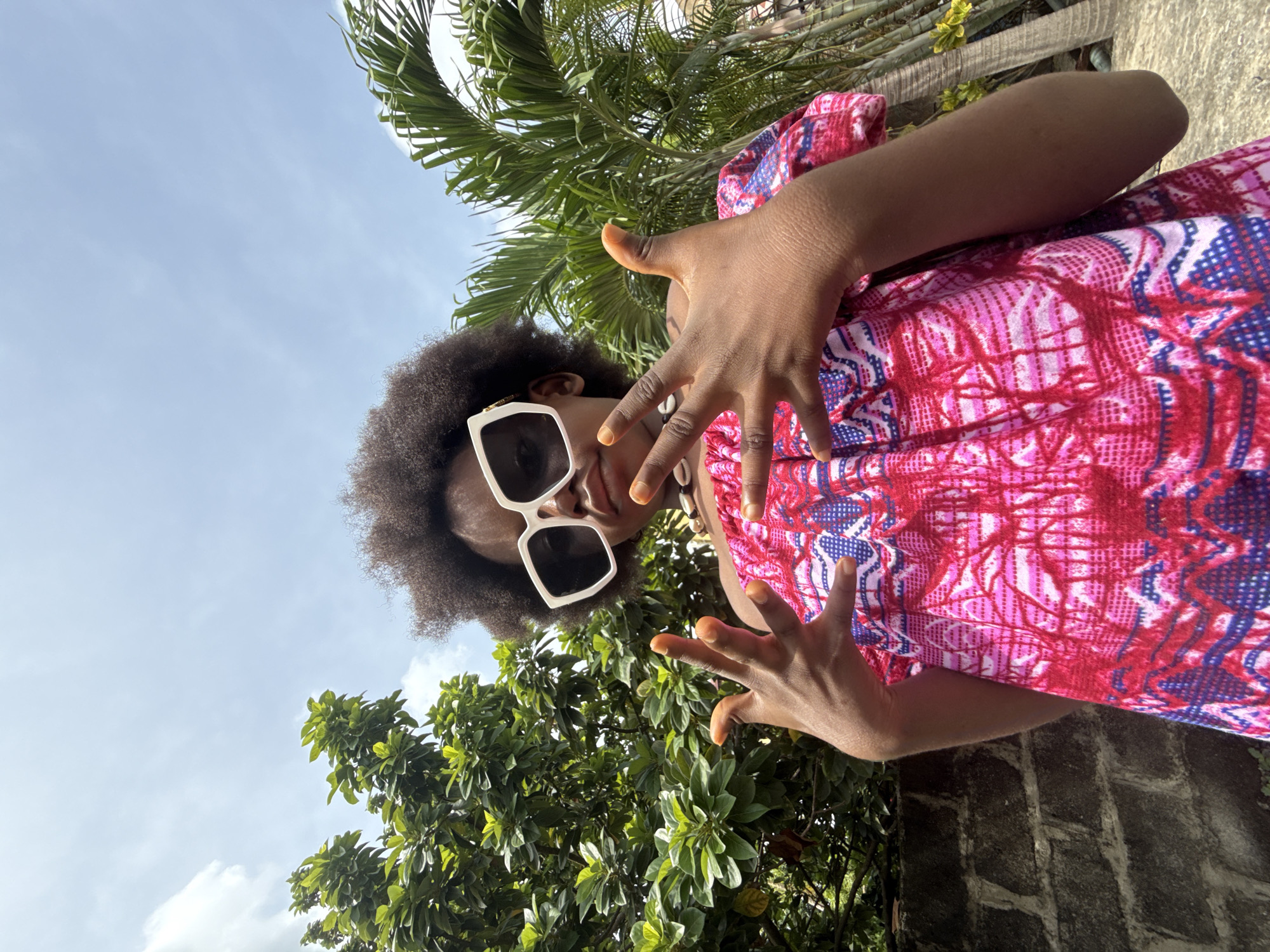 A low-angle shot depicts a person with brown skin wearing a pink and red patterned garment and large, square, white sunglasses, standing outside in Ado, Nigeria. The person's hair is a dark afro. They extend both hands towards the camera. The background features a blue sky with a palm tree and lush green foliage. A brick wall is visible in the bottom right corner.