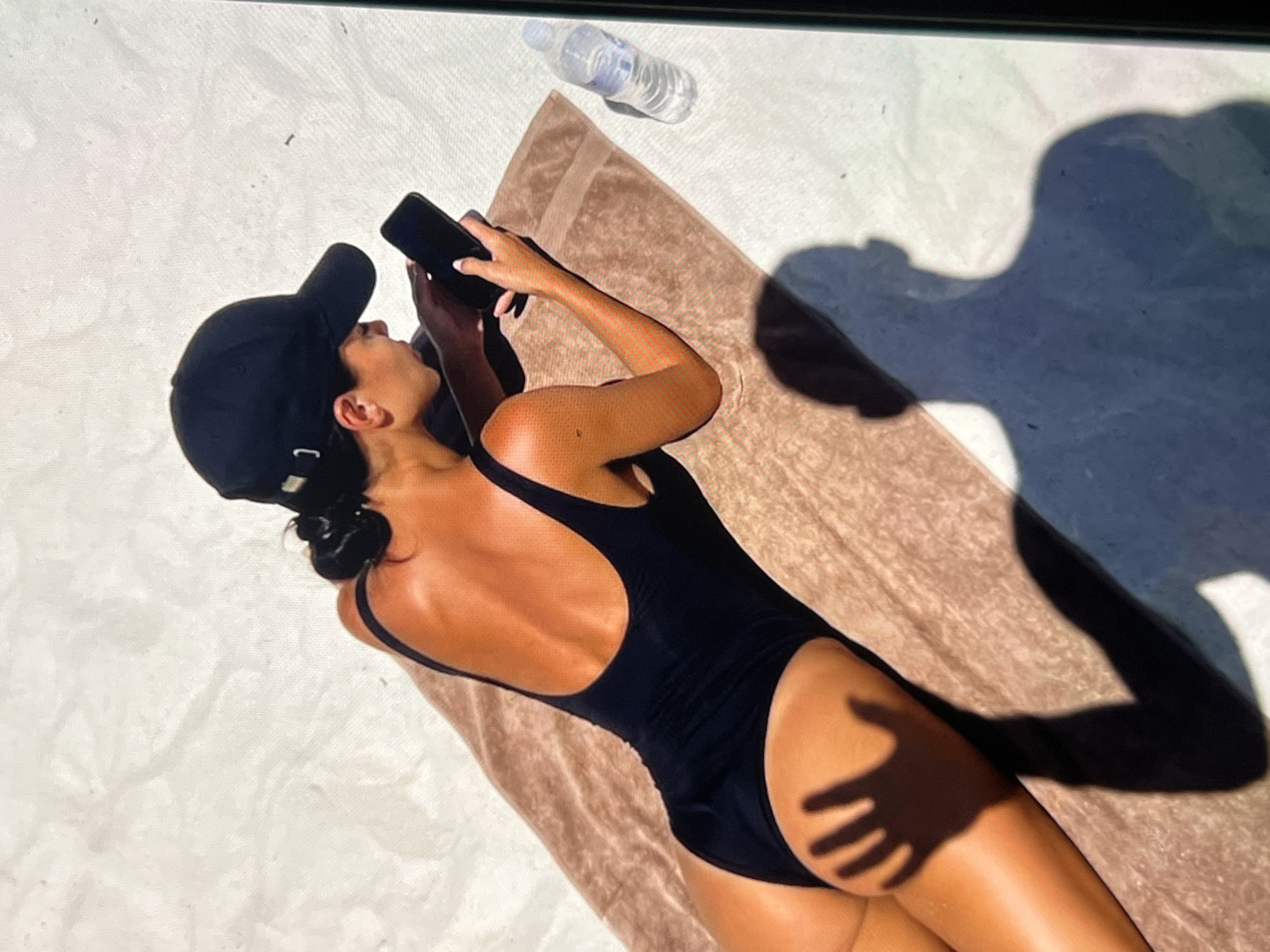A person wearing a black swimsuit and a black baseball cap lies on a tan towel on a sandy surface, holding a smartphone. A plastic bottle of water is to the right of the towel. The person is seen from a high angle, with shadows cast on the sand. The location is not specified as being in Montfermeil, France.