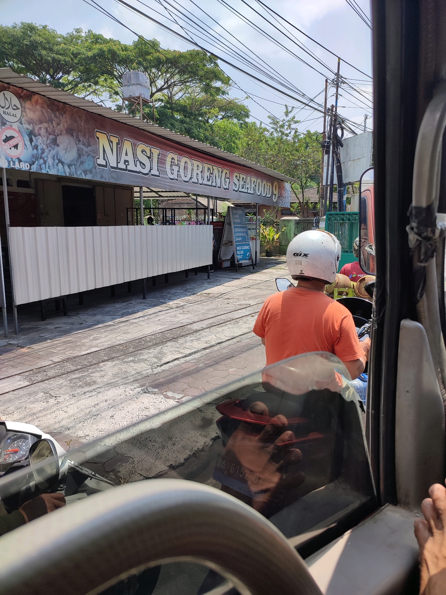 In the city of Magelang, Indonesia, a medium shot captures a street scene with a focus on a motorcycle rider and a food stall. The rider, wearing an orange shirt and a white helmet, is positioned in the foreground, facing away from the camera. The motorcycle itself is partially visible, with its handlebars and dashboard in clear view.
Behind the rider, a food stall with a banner displaying "NASI GORENG SEAFOOD 9" is prominent. The banner also features a "HALAL" logo and a "NO PORK LARD" symbol. The stall has a long, white, corrugated metal barrier in front of it, supported by dark legs. Adjacent to the stall are some plants and a green fence.
The environment suggests a sunny day, with a clear sky and power lines crisscrossing overhead. The ground is paved and shows signs of wear. The reflection on the motorcycle's windshield reveals a glimpse of the person taking the photo and a hand resting on the dashboard. The overall scene depicts a typical street view in an Indonesian city, focusing on local commerce and daily life.