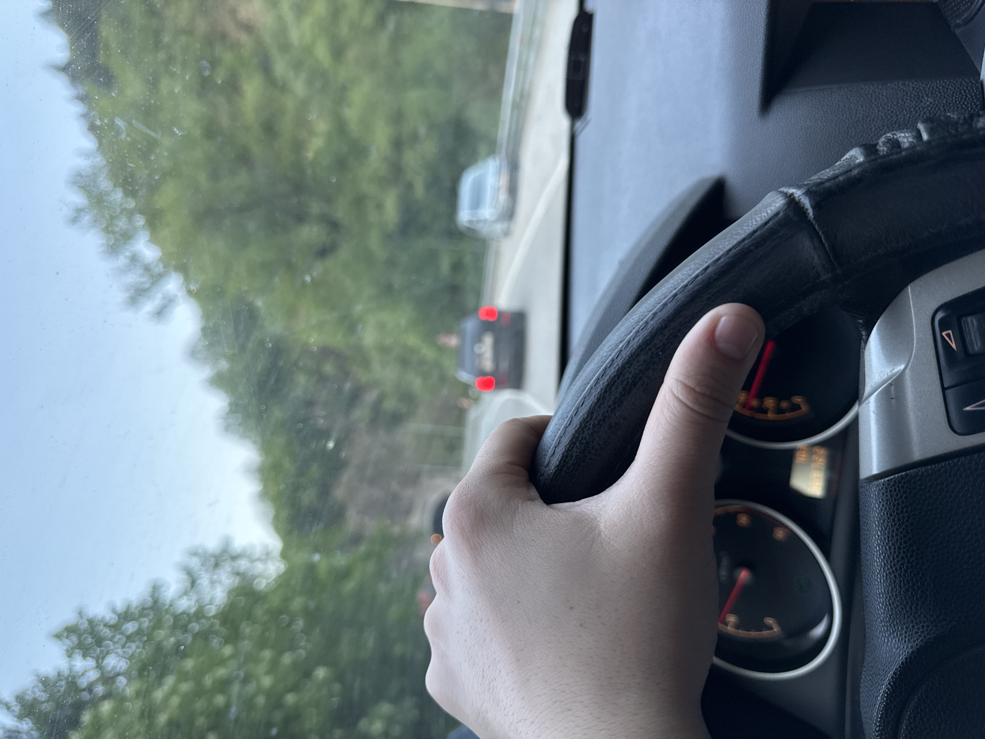 The image depicts the interior of a car, with a driver holding the steering wheel. Through the windshield, another vehicle can be seen on a road surrounded by trees. The speedometer is visible to the right of the steering wheel. The location is possibly near Бачковски манастир, Bulgaria, based on the surrounding environment.