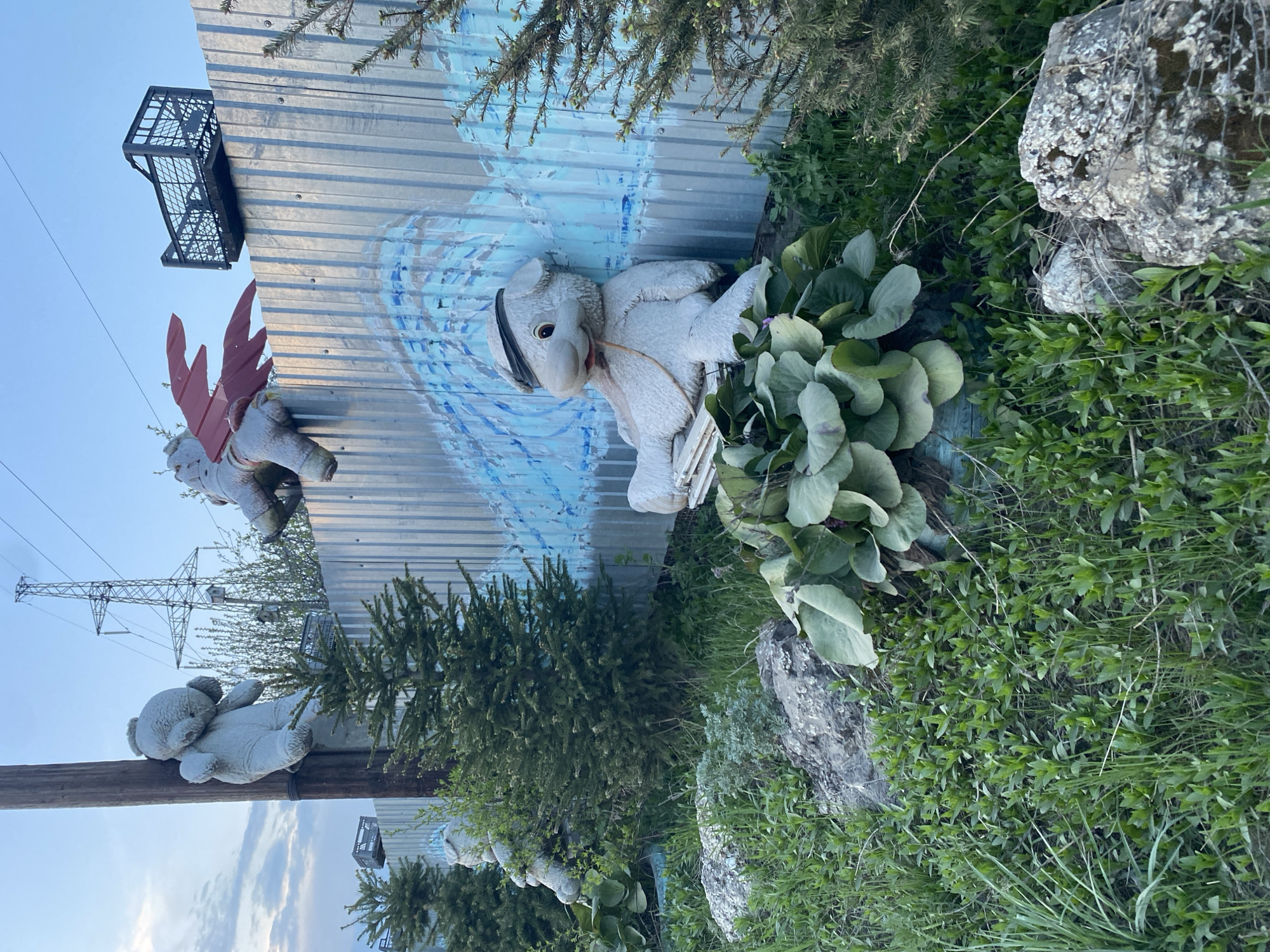 The image shows a collection of outdoor art installations in the city of Chaikovsky, Russia. Three teddy bear statues are the primary subjects. One is attached to a utility pole, and another appears to have red wings. A third teddy bear is resting against a metal wall painted with blue and white patterns, resembling waves. The background includes greenery, trees, rocks, and a utility pole. A black crate is attached to the top of the metal wall.
