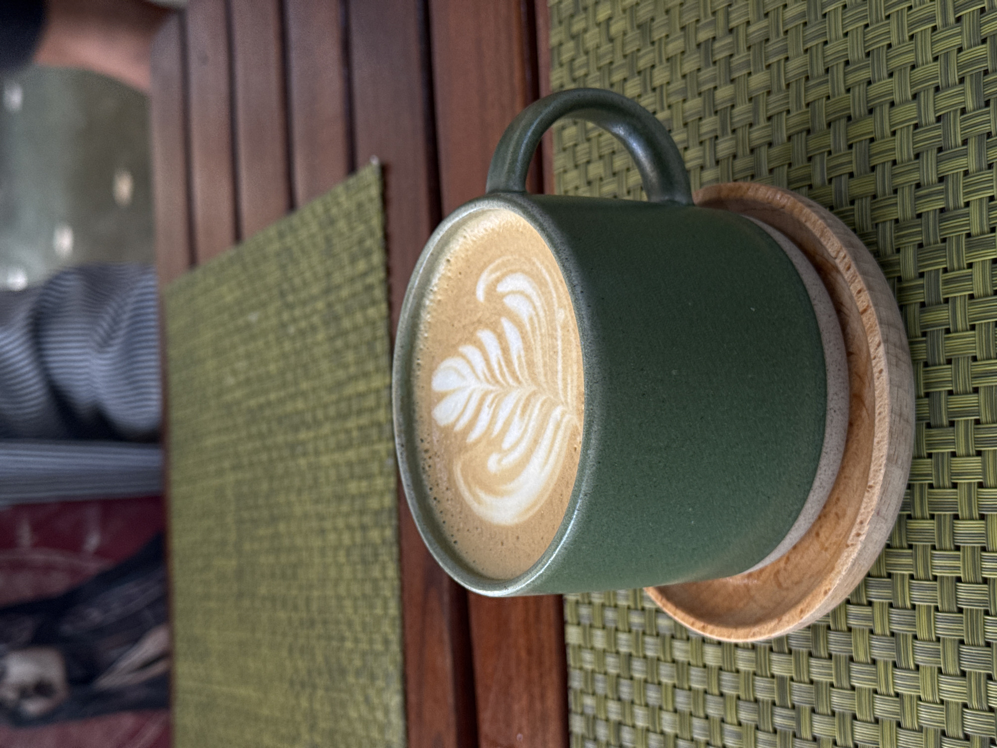 A close-up, eye-level shot focuses on a green ceramic mug of latte art, placed on a small wooden saucer. The mug is set on a green woven placemat atop a slatted wooden table. In the blurred background, the upper body of a person wearing a striped shirt is visible, along with another indistinct figure. The setting appears to be an outdoor cafe in the city of Ko Samui District, Thailand.