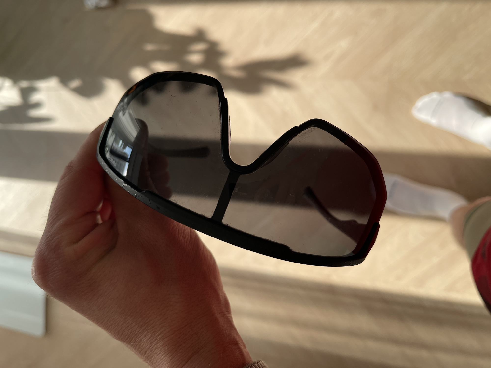 A person is holding a pair of dark, sport-style sunglasses in their hand. The background is a light-colored wooden floor. A leg wearing a white sock is visible in the upper right of the frame. No features in the image suggest the location is Izhevsk, Russia.