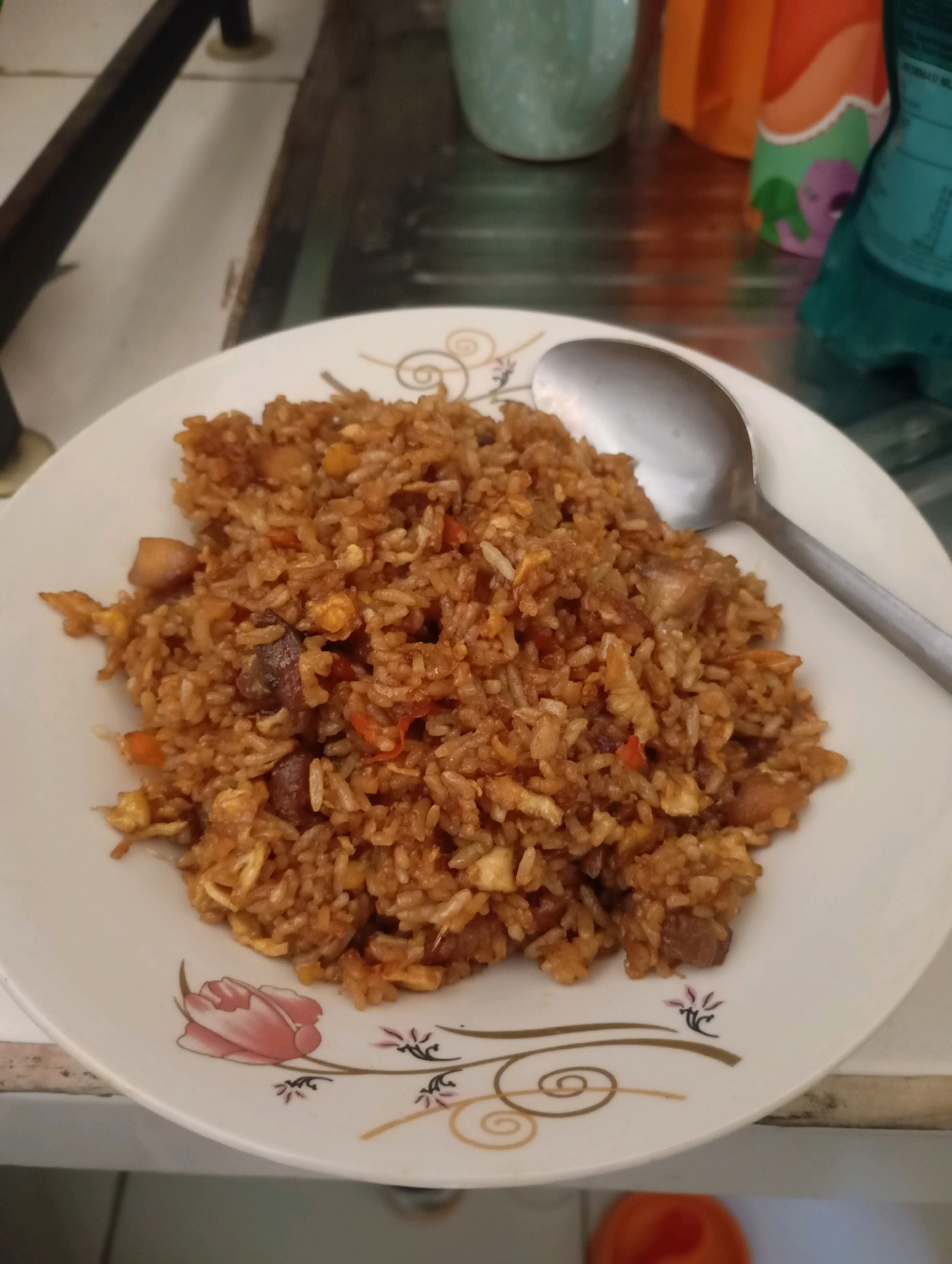 A close-up, eye-level shot shows a white plate filled with a generous portion of fried rice. The rice is a rich brown color, indicating it has been stir-fried with soy sauce and other seasonings. Mixed throughout the rice are visible pieces of protein, likely chicken or pork, along with bits of scrambled egg and possibly chopped vegetables such as carrots. A silver spoon rests on the upper right side of the plate, partially submerged in the rice, suggesting it is ready to be eaten. The plate itself has a decorative rim featuring a floral pattern in shades of pink and brown, and a swirling, golden design. The background is blurred but appears to be a kitchen or dining area, with hints of colorful bottles and a countertop. The image is lit from above, casting a slight shadow on the lower part of the plate. The scene is set in Babelan, Indonesia.