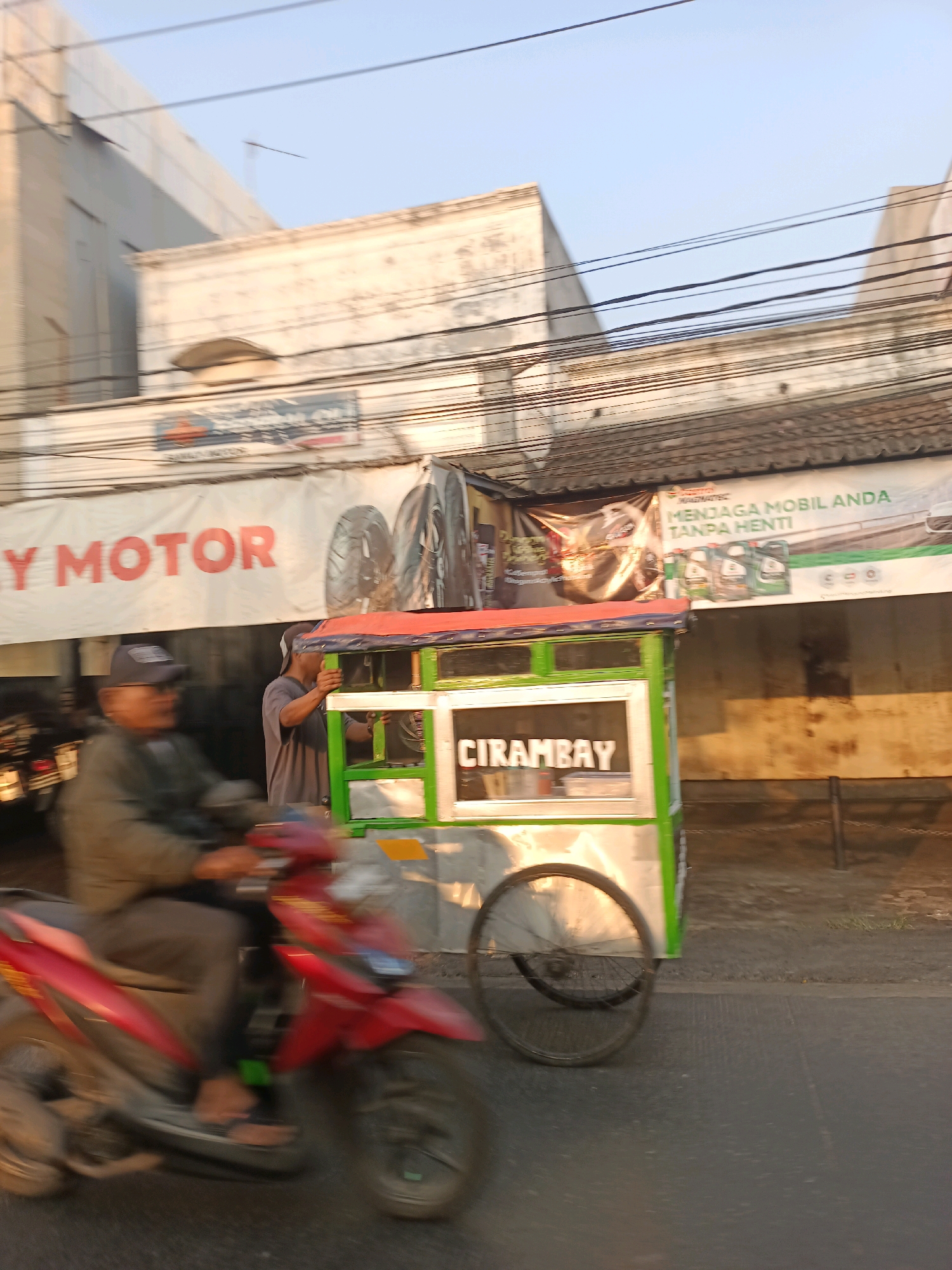 The image captures a street scene in Karawang, Indonesia, with a focus on a food cart and a passing motorcycle. The food cart, painted green and silver, is positioned on the right side of the frame. It appears to be a mobile vendor, with a wheel visible on its left side. The word "CIRAMBAY" is clearly visible on a sign on the cart. Two individuals are near the cart; one is partially obscured, appearing to interact with the cart, while another man in a grey shirt is standing behind the cart, possibly attending to it. In the foreground, a red motorcycle is in motion, with its rider, a man wearing a dark jacket and a cap, blurred due to the movement. The background features buildings and overhead wires, indicating an urban setting. A banner with text in Indonesian, including "MENJAGA MOBIL ANDA TANPA HENTI," is displayed on the building behind the food cart. The lighting suggests it is either early morning or late afternoon.
