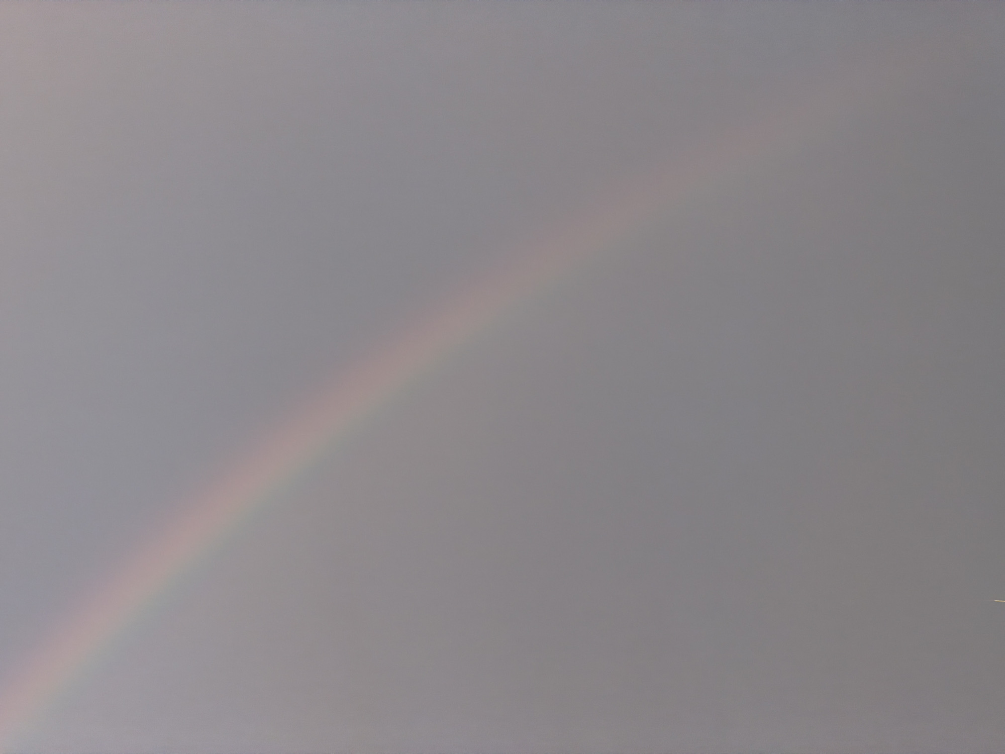 The image displays a subtle rainbow arc against a muted, overcast sky. The sky occupies most of the frame and is a uniform grey, with no discernible clouds or celestial bodies. The rainbow is faint, appearing as a soft band of pastel colors, predominantly pinks and pale yellows, with a hint of green. It stretches diagonally from the upper left to the lower right of the image. The lower edge of the image shows a sliver of what might be a distant structure, possibly a building or a silhouette, appearing as a dark, indistinct line. The scene lacks any clear indicators of a specific location within Kazan, Russia, or any identifiable people, subjects, or actions.