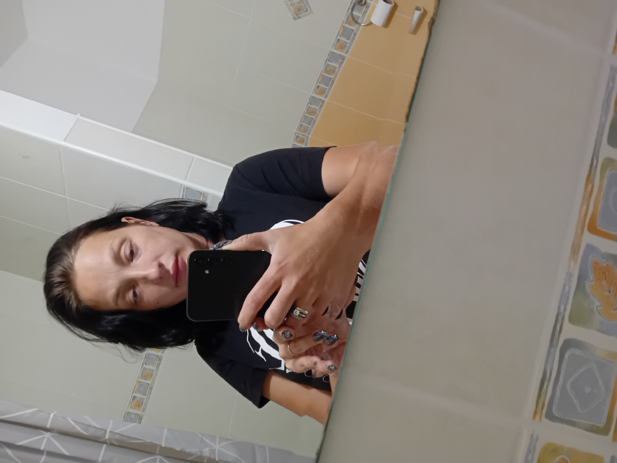 A woman with dark hair, wearing a black t-shirt, takes a mirror selfie in a bathroom. She is holding a black smartphone in her right hand. Her left hand is visible, showing painted nails and rings. The bathroom features tiled walls with a patterned border and a light-colored shower curtain. A toilet paper roll holder is visible on the wall. The scene is likely indoors.