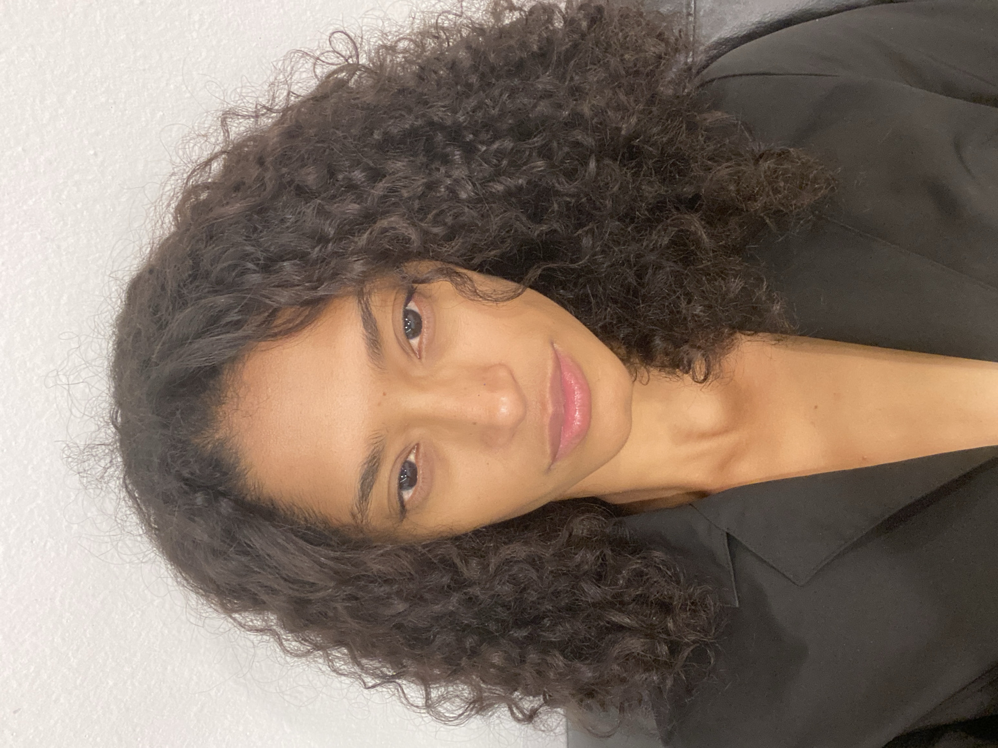 The image is a portrait of a woman, oriented such that her head is on the left and her body extends towards the right. She has voluminous, dark, tightly curled hair that frames her face and shoulders. Her skin is clear, and she has full, subtly pink-tinted lips. Her dark eyes are open and directed towards the viewer, conveying a calm and gentle expression, possibly with a faint smile. She is wearing a black garment, likely a jacket or top, which is visible on her right side, partially covering her chest and shoulder.
The background consists primarily of a plain, light-colored (possibly white or off-white) wall. A darker, indeterminate surface is visible behind her upper right shoulder. The lighting is soft and even, suggesting an indoor setting during the daytime. There is no visible text in the image. While the location context provided is São Paulo, Brazil, there are no specific visual cues within the image itself to confirm this.